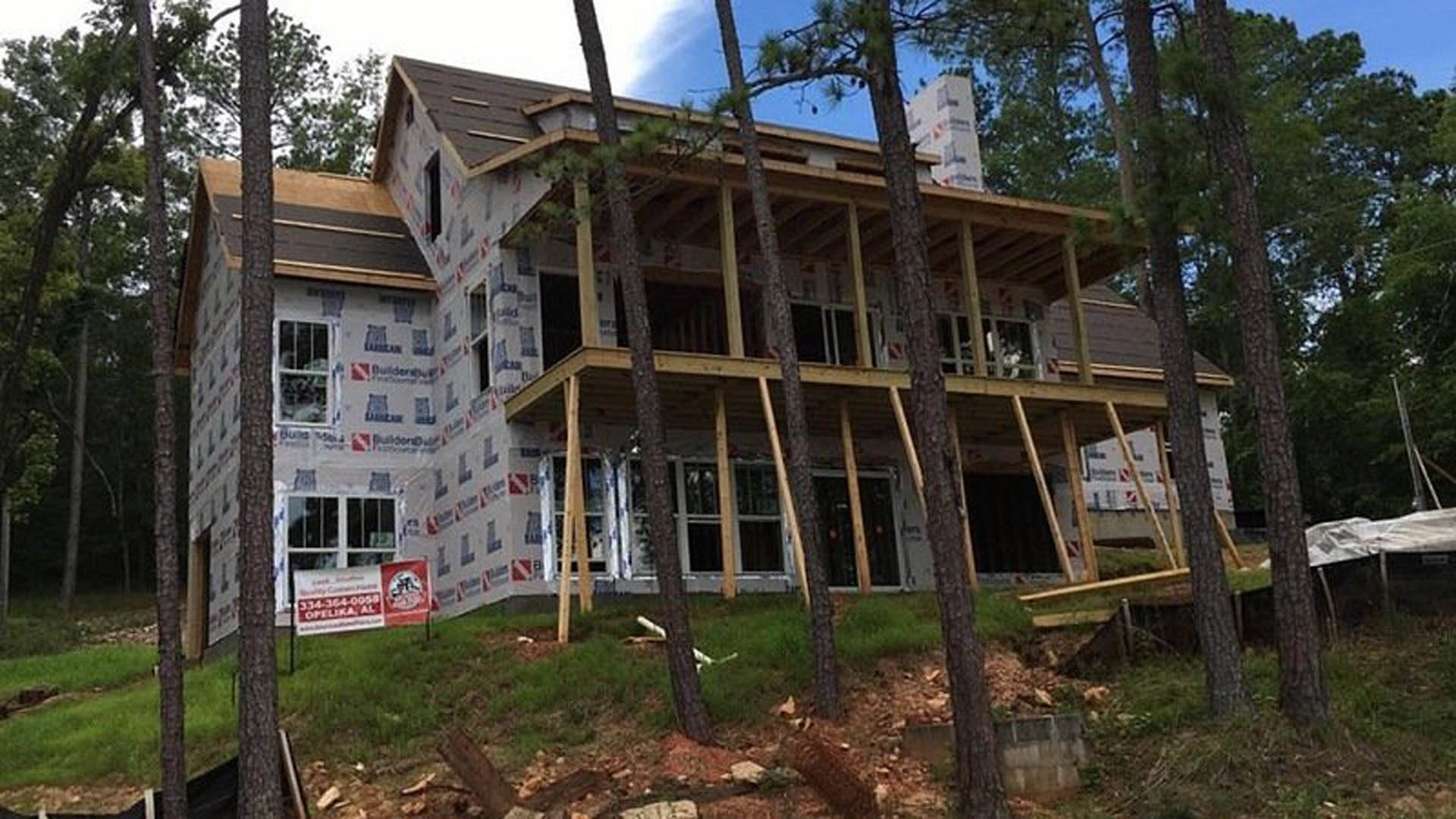 Framed house under construction surrounded by mature trees, exposed wood beams, window with metal bars, grassy yard with tree stumps, red construction sign and black utility boxes
