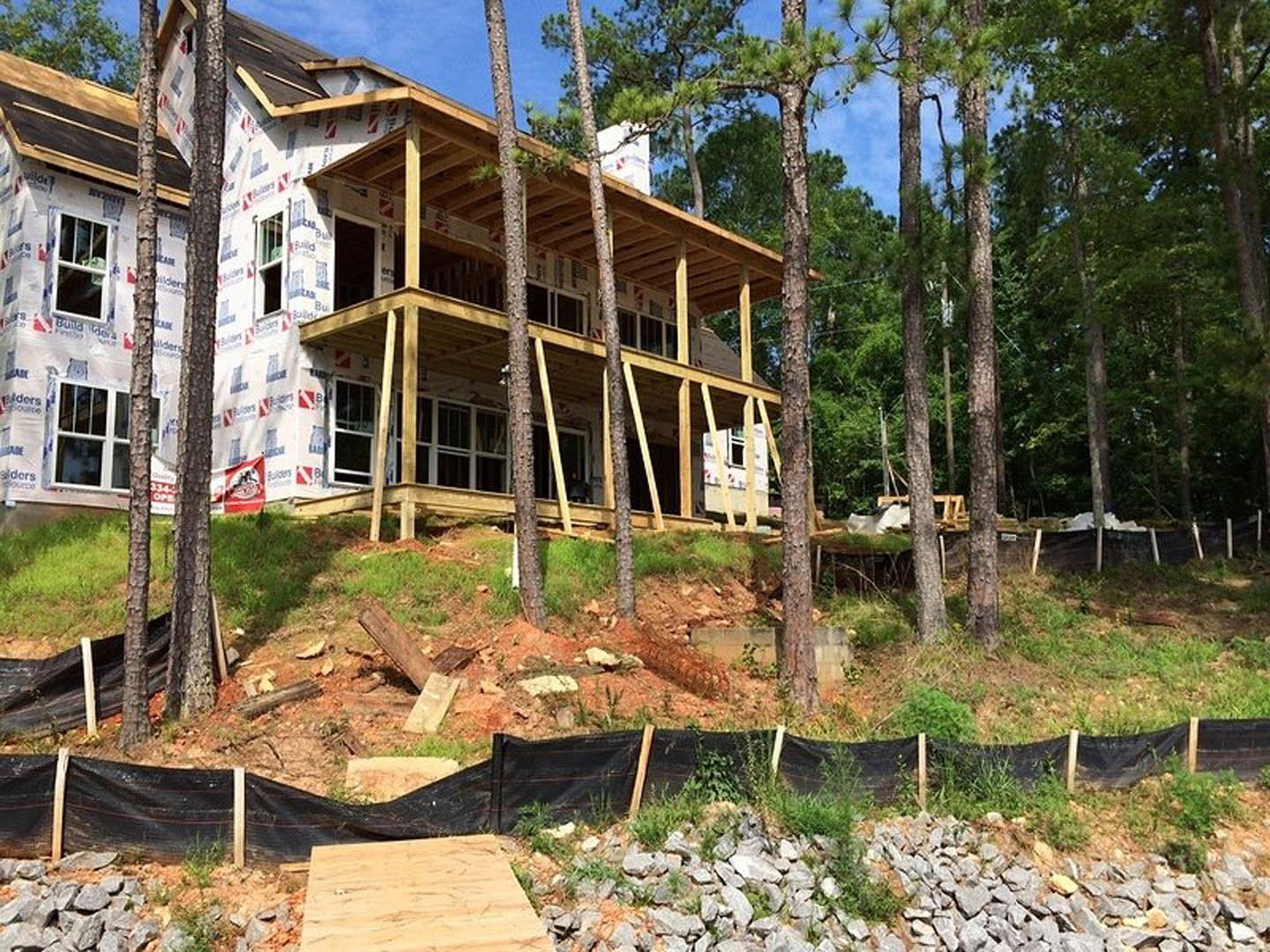 Framed house under construction surrounded by mature trees, exposed plywood sheathing, black rectangular window openings, red and white construction flag, wooden surfaces, black