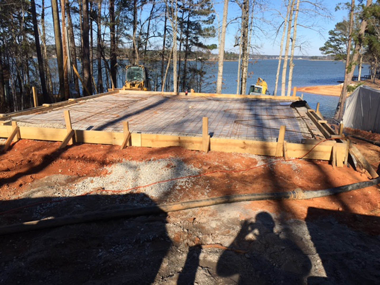 Wooden foundation and beams at residential construction site near a pond, surrounded by trees, with construction vehicles and equipment visible