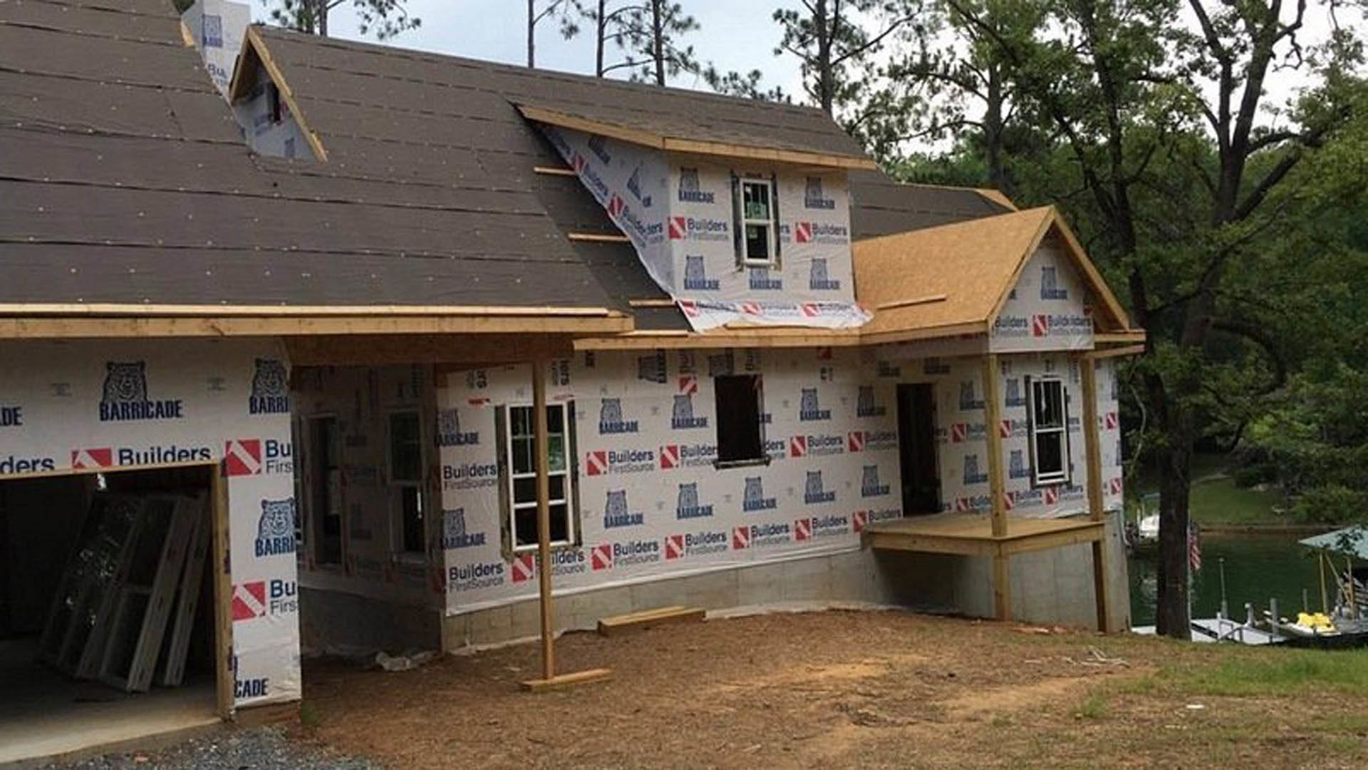 Two-story house under construction with gray siding, shingled roof, covered front porch, and large windows, surrounded by dirt yard and mature trees