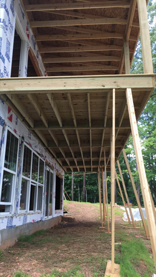 Two-story house framed with exposed wood beams and planks, partially built deck overlooking grassy yard, construction materials scattered near dirt path, trees and plants in