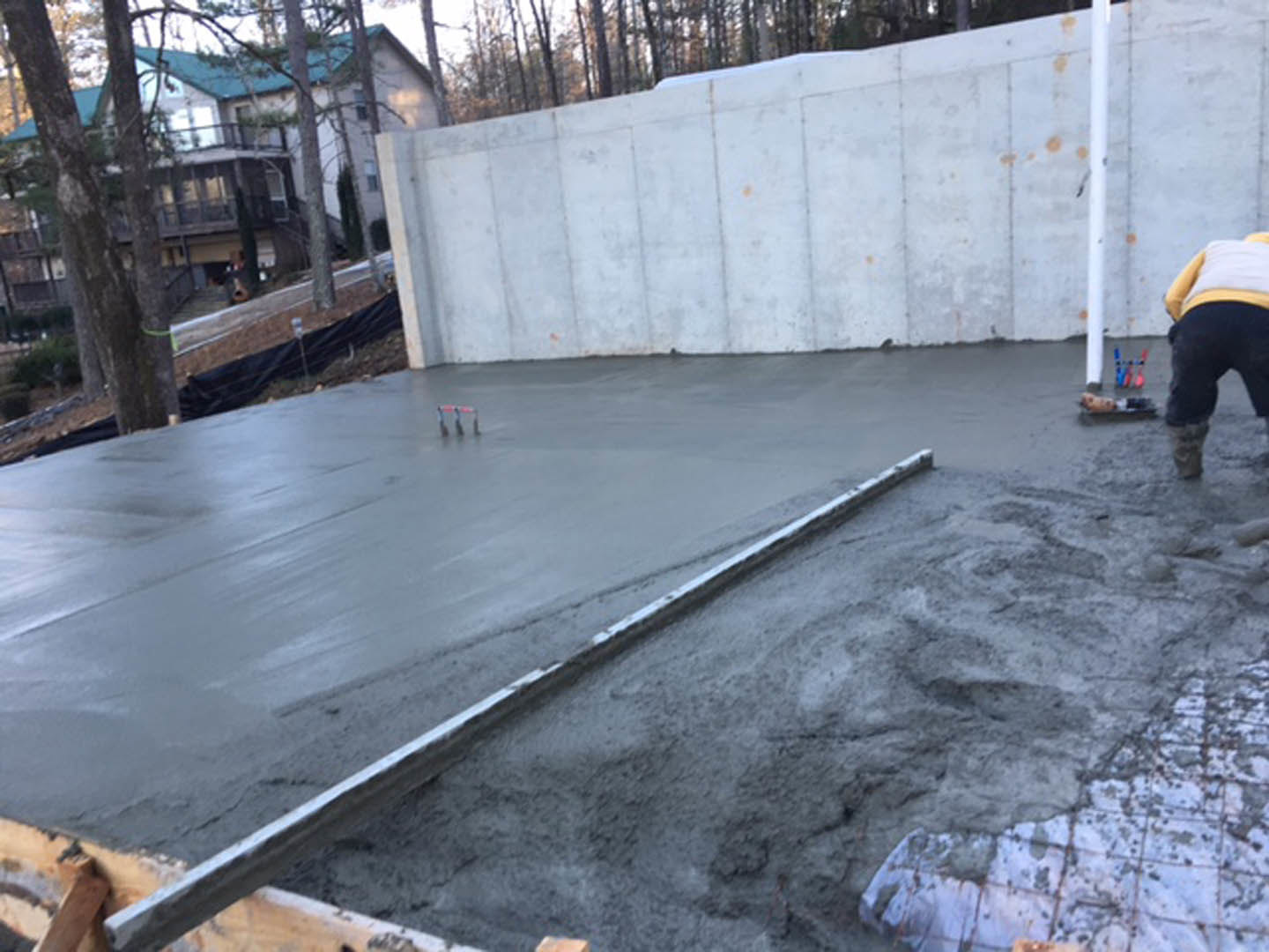 Freshly poured concrete slab with wooden framing, worker in black pants smoothing surface, white wall and trees in background, blurry foot visible in foreground