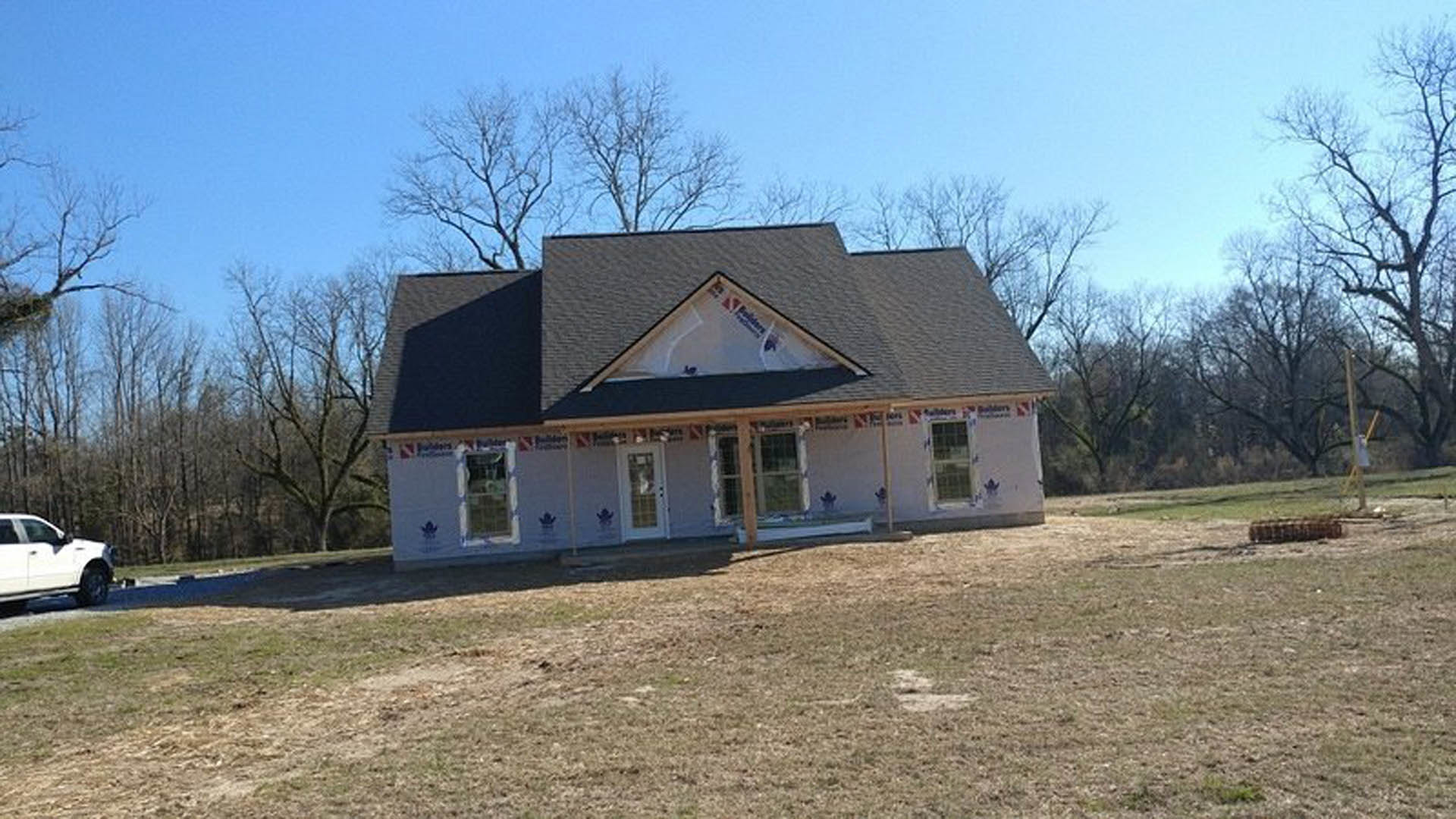 Covered porch with white columns on a newly constructed house, grassy front yard, white car parked on the road, large windows with glass panes, red sign visible in one window.