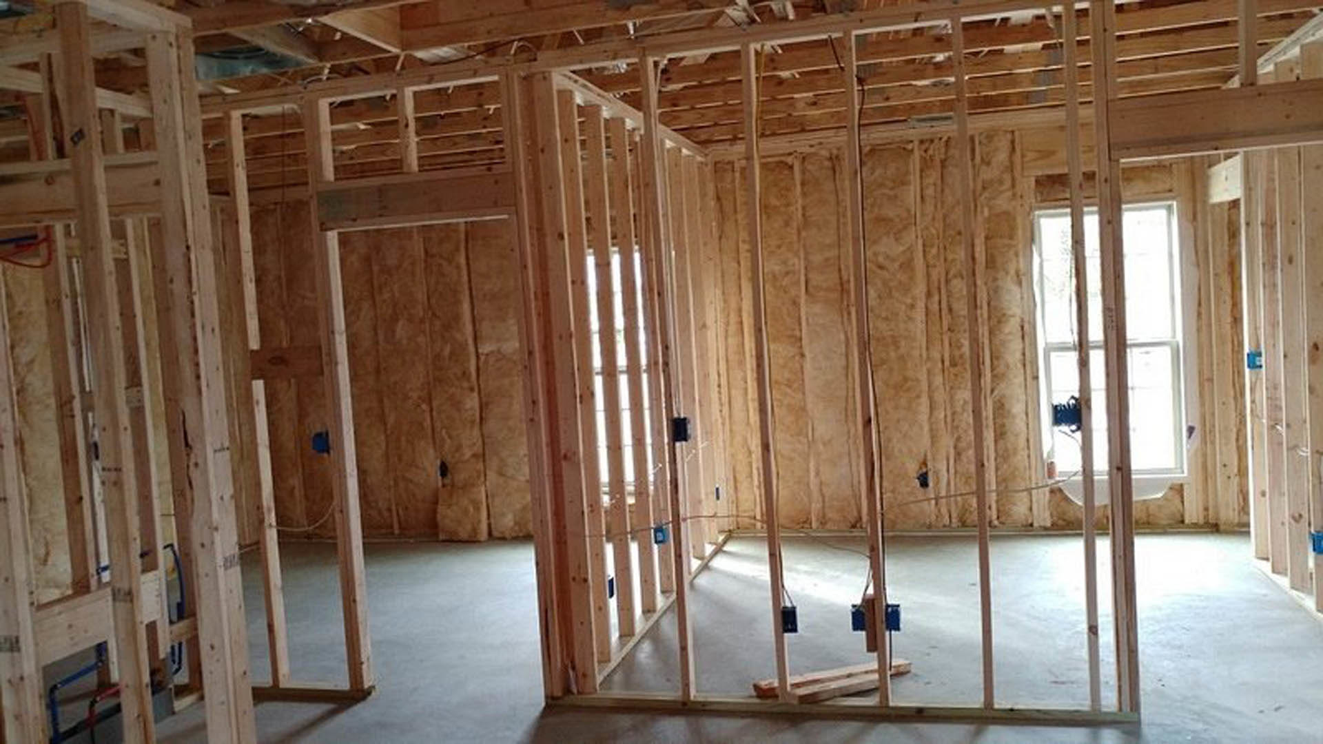 Exposed wood framing and beams in an unfinished room, with construction materials, insulation, and a ladder visible on the floor.