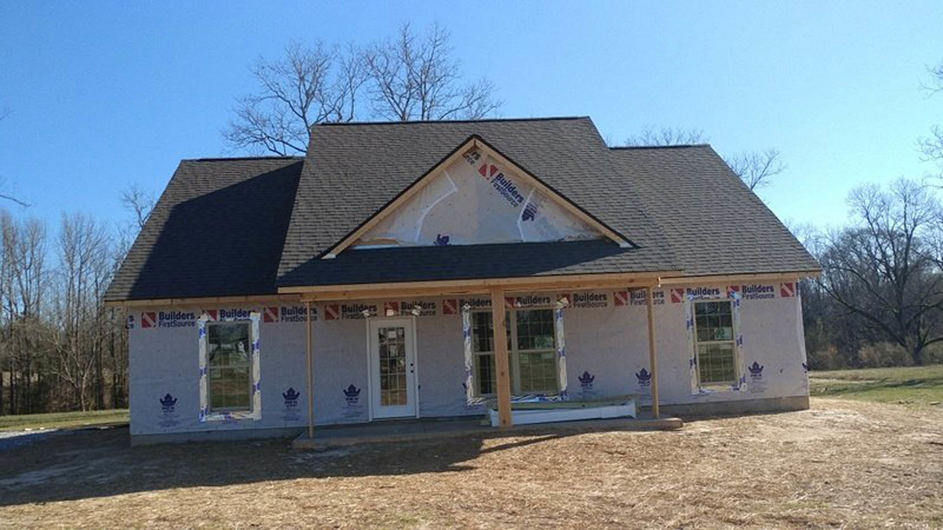 Modern cottage-style house under construction featuring completed shingle roof, exposed wooden framing, large windows, leafless tree in foreground, and white surface with builder’s