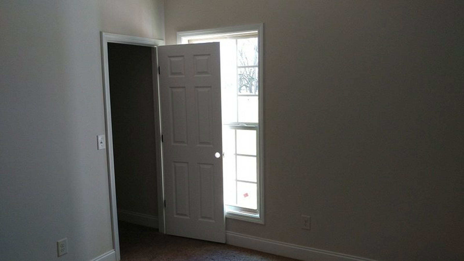 Open white door with silver handle in a bright room, wall outlet visible near baseboard, large window lets in natural light, neutral painted walls and hardwood flooring