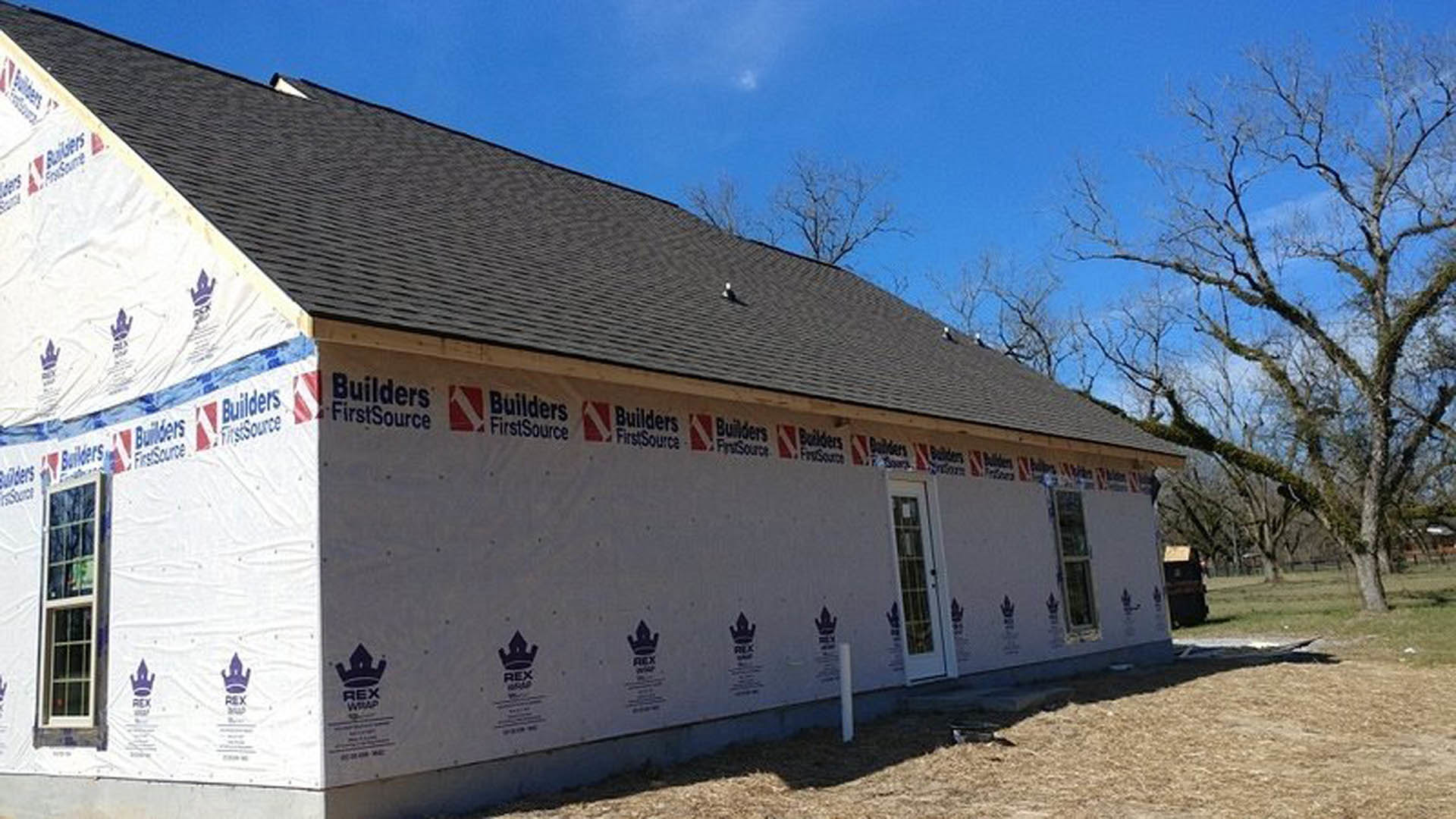 Two-story house under construction with exposed framing, white signs attached to exterior walls, leafless tree nearby, barred window visible, cloudy sky overhead