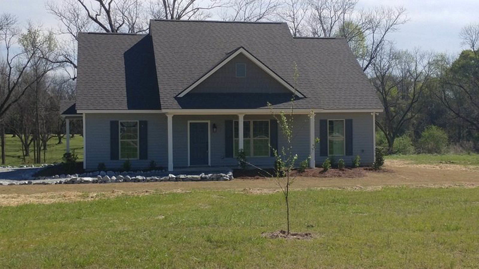 Two-story house with white siding, covered front porch, large windows, manicured lawn, and mature trees surrounding the property