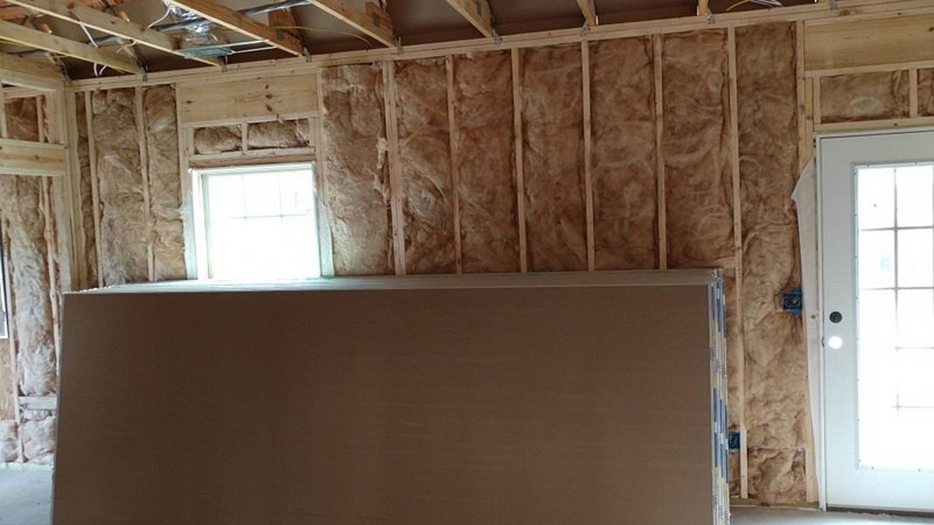 Sunlight streaming through a rectangular window onto plaster walls, exposed wooden beams and plywood ceiling, white door with glass inset visible
