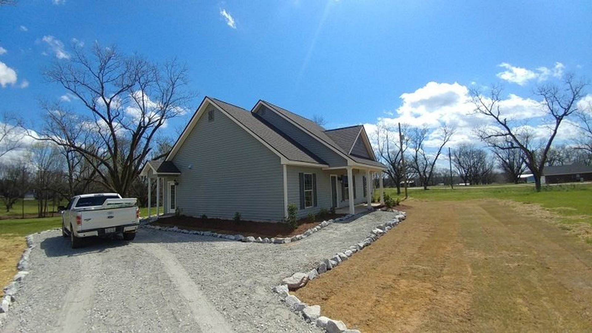 Two-story house with gabled roof, large windows, and white pickup truck parked on driveway; manicured lawn, mature tree, and blue sky with scattered clouds