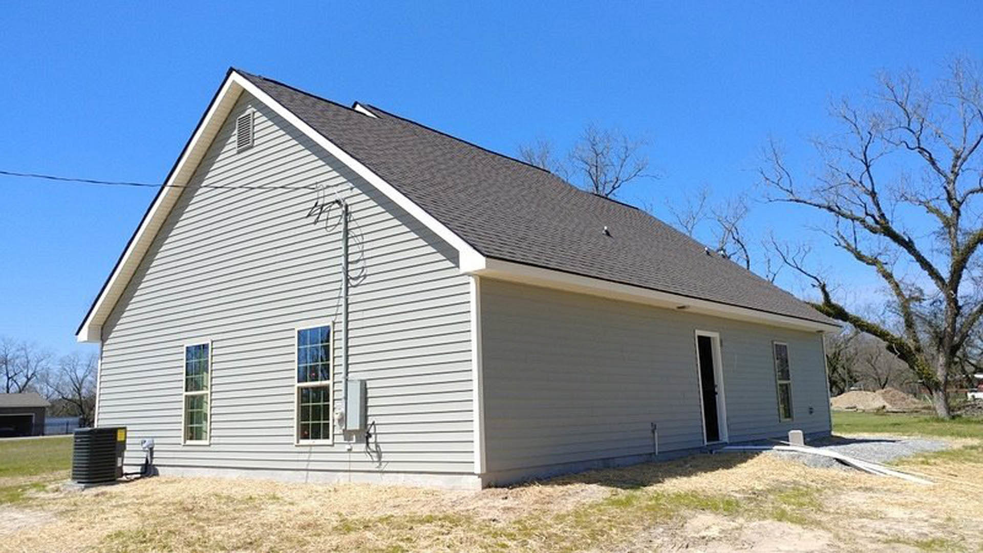 Side view of a cottage-style home with light-colored siding, minimal windows with white frames, leafless tree in foreground, and a dark entry door