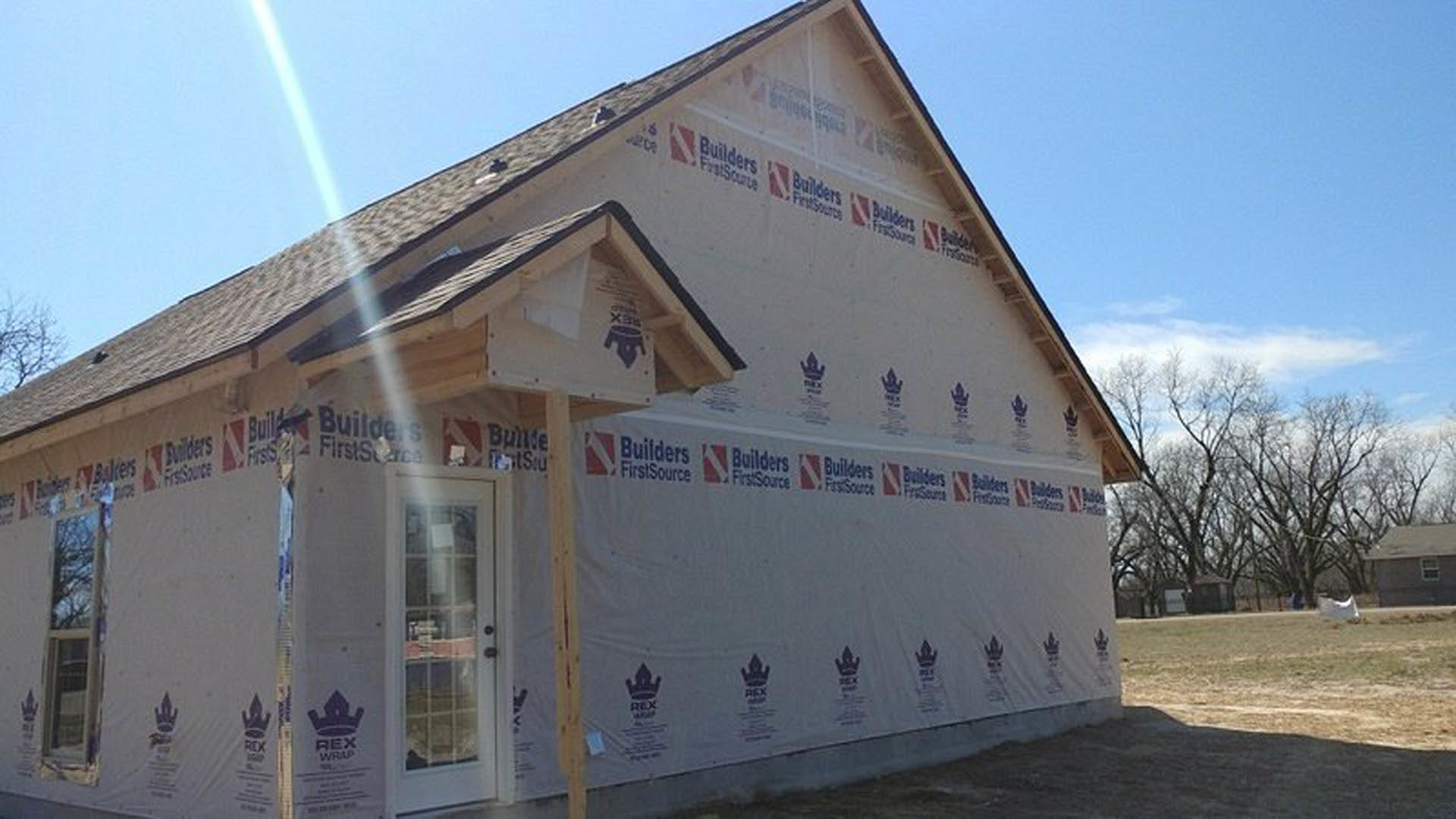 Two-story house under construction wrapped in white protective sheeting, surrounded by trees and neighboring homes, cloudy sky overhead