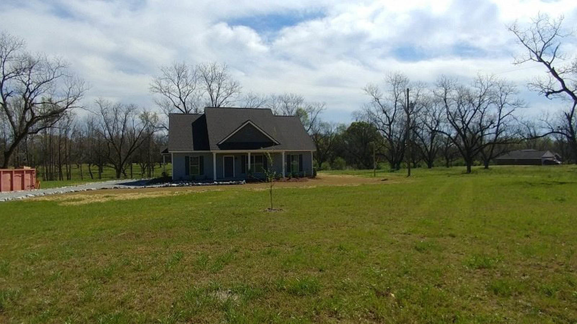Modern home with black roof surrounded by grassy field, mature tree in front yard, large windows, and scattered clouds in blue sky