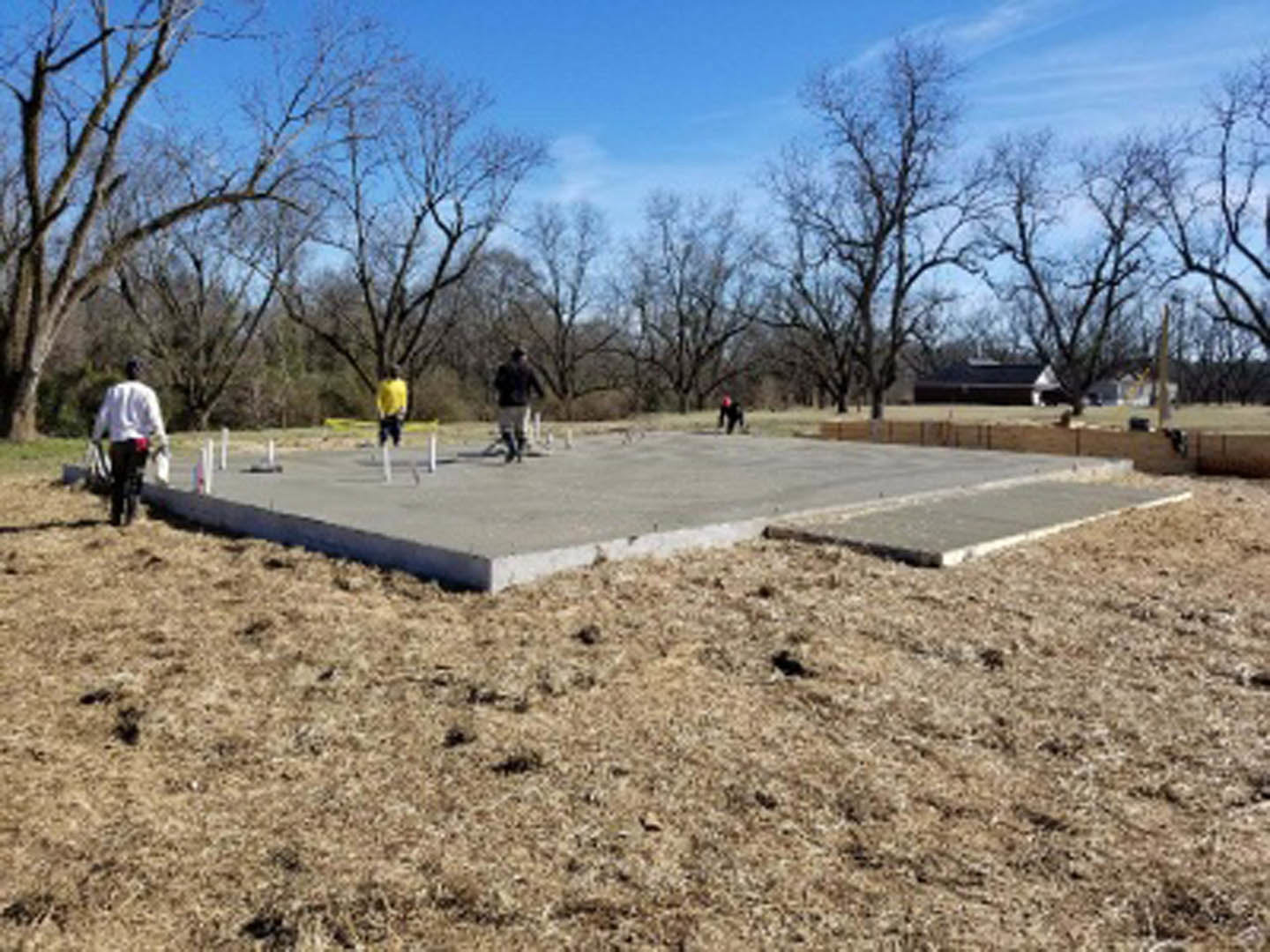 Group of people gathered on unfinished concrete foundation surrounded by trees and blue sky