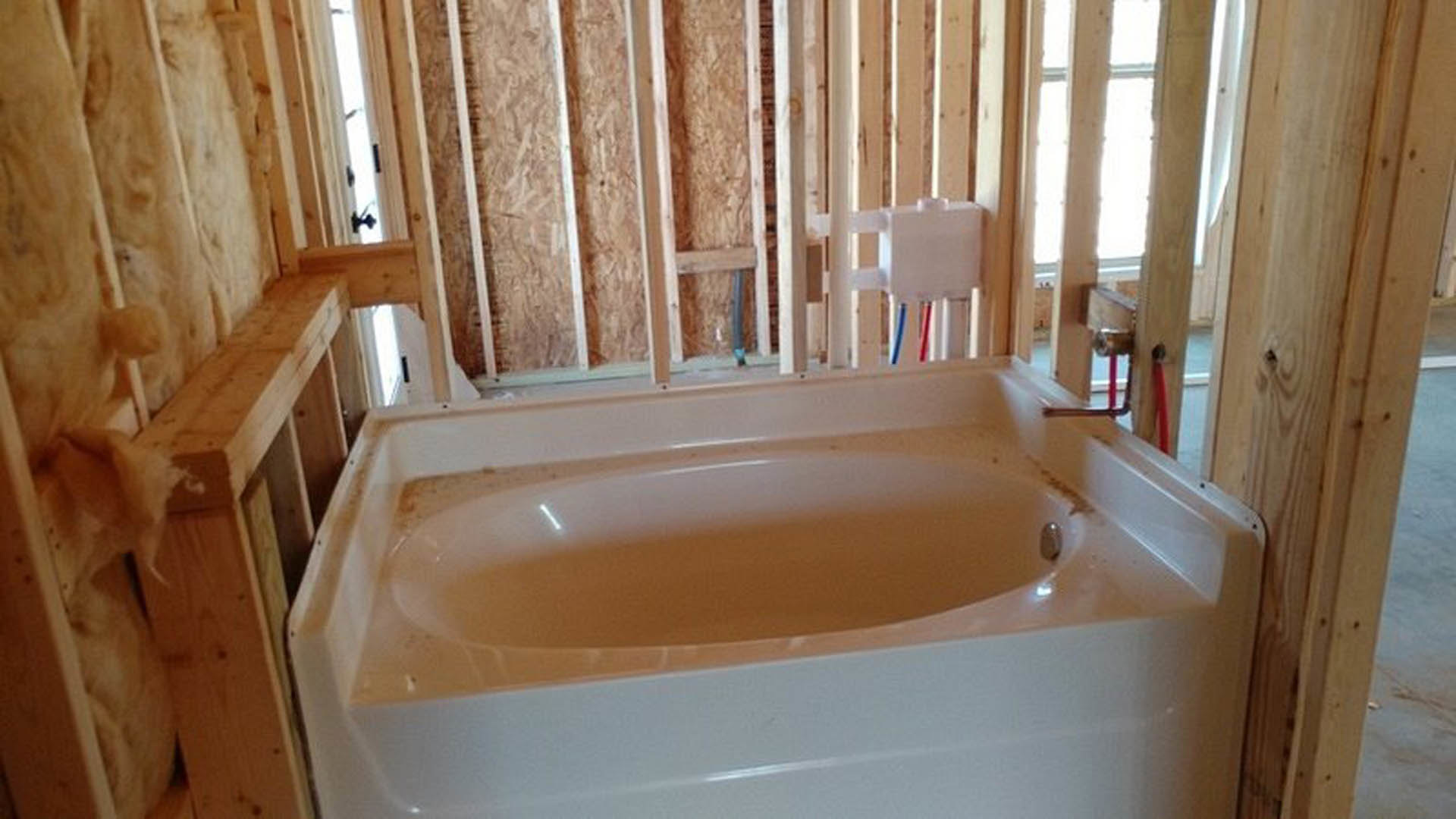 White bathtub installed in unfinished bathroom with exposed wood framing, visible plumbing pipes, and construction materials scattered nearby.