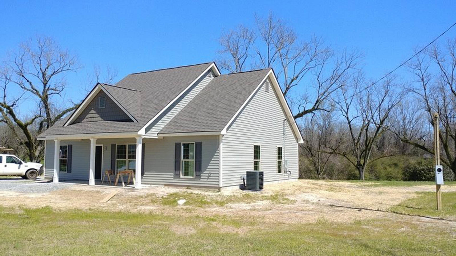 Two-story house with covered porch, white-framed windows, expansive green lawn, mature trees in background, white pickup truck parked on road, grey utility box near driveway