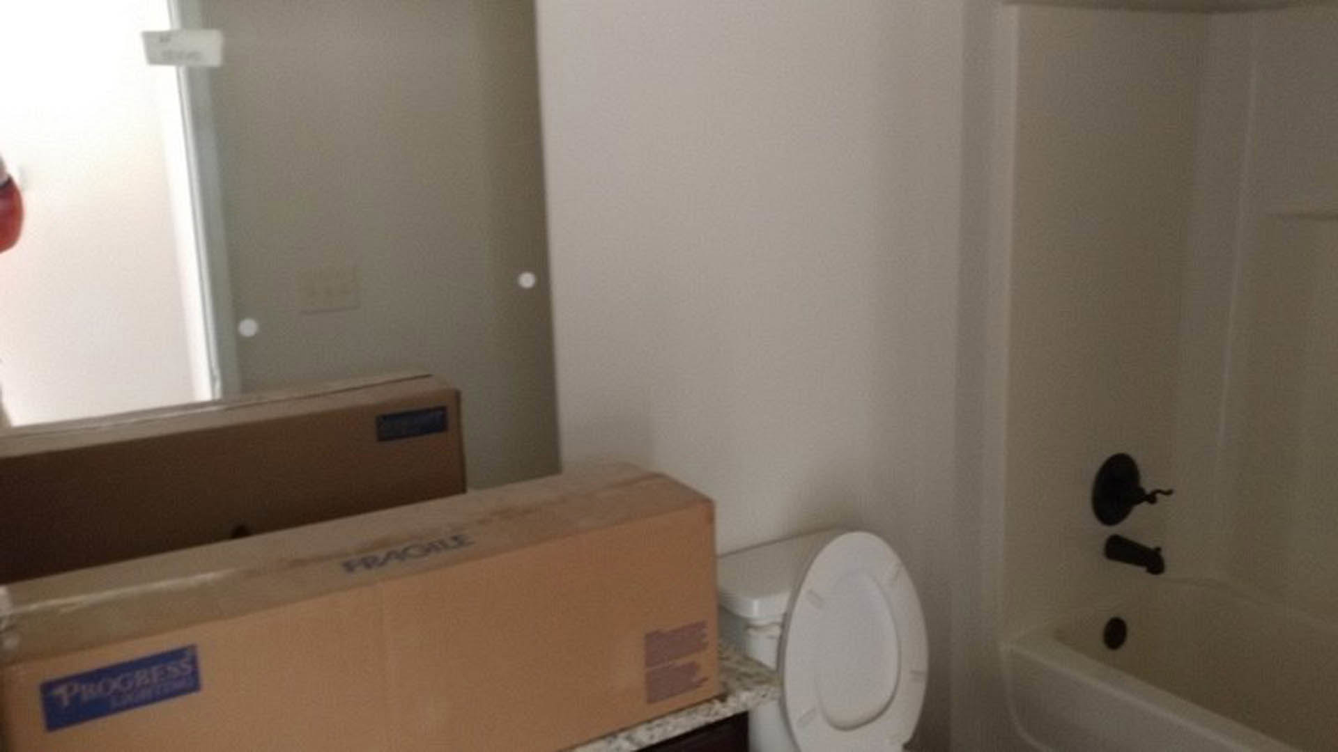 White toilet with lid open beside a rectangular box on tiled bathroom floor, white walls and partial view of bathtub and mirror