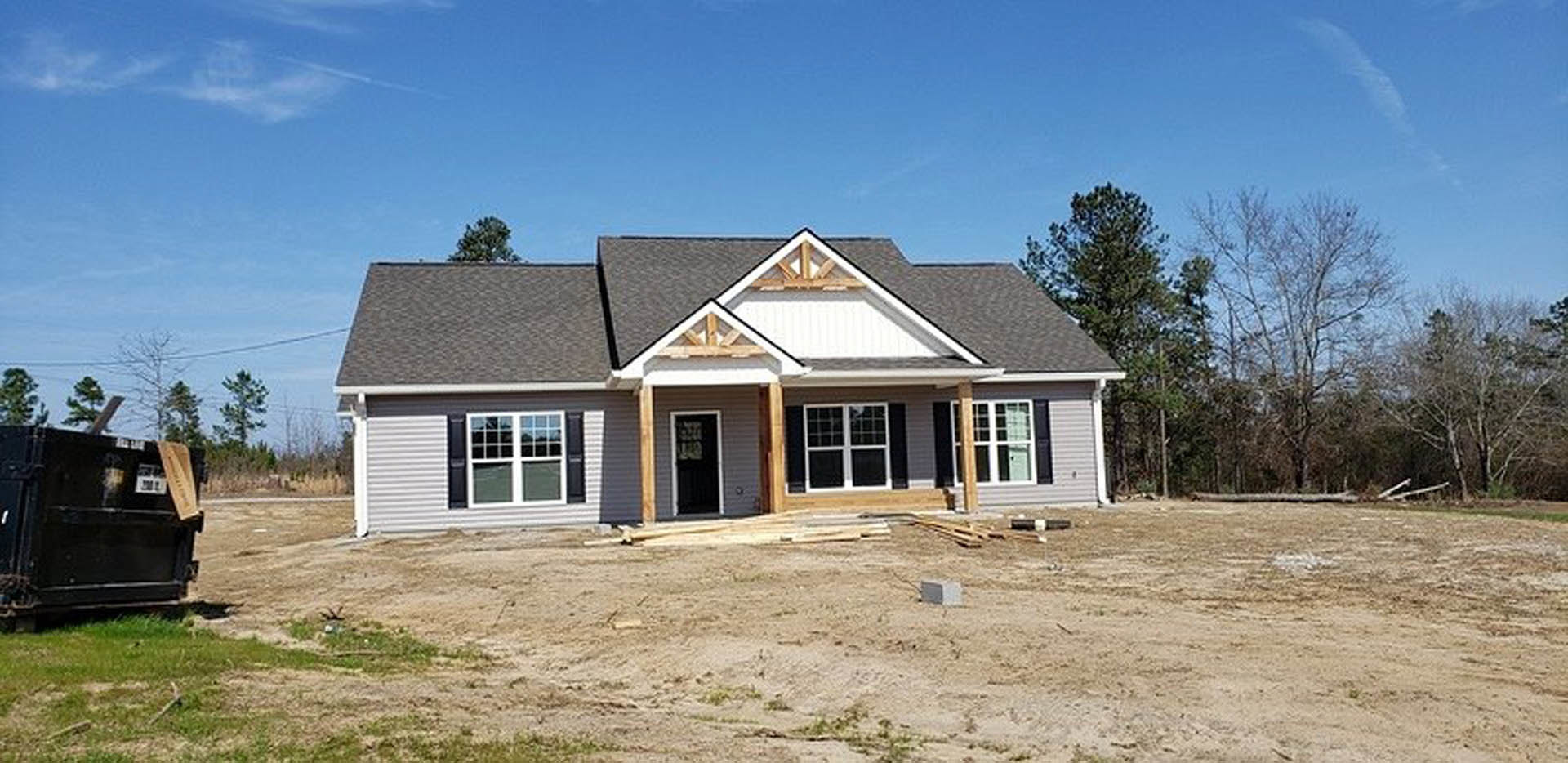 Partially built house with exposed framing and white window trim, surrounded by dirt field; black waste container with signage and construction materials nearby; cloudy sky and