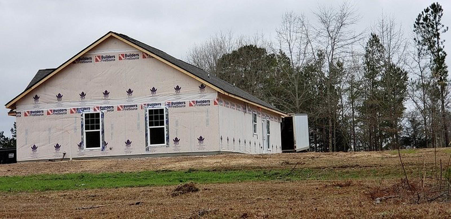 Partially built farmhouse with exposed framing, unfinished exterior walls, and surrounding grassy lot bordered by mature trees under a cloudy sky