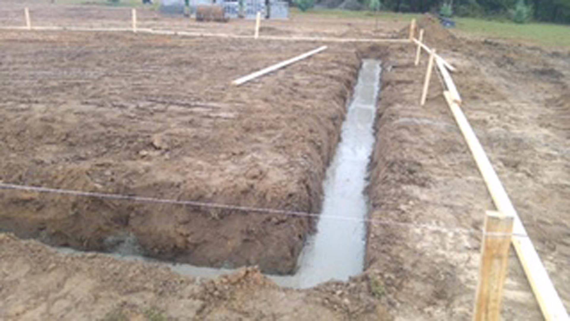 Narrow dirt trench running through grassy yard with exposed soil and wooden beam along foundation