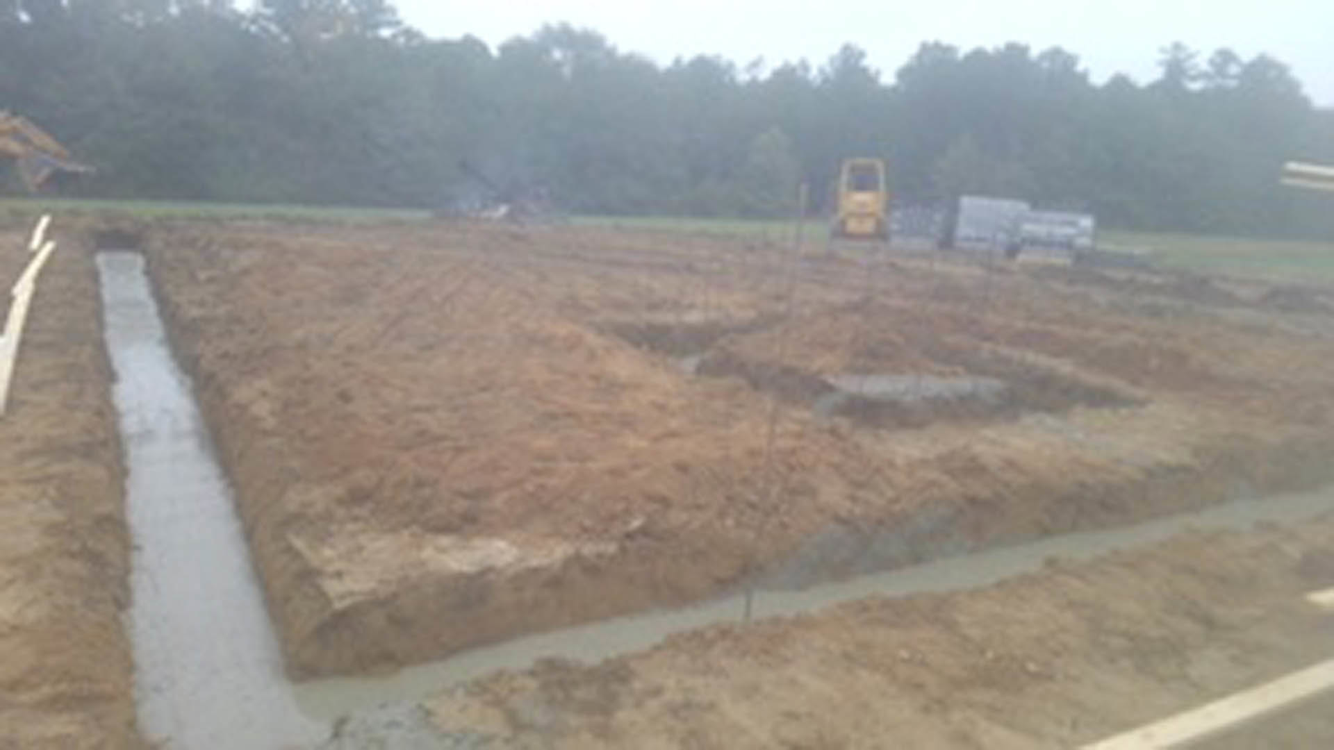 Construction site with exposed soil, metal poles, and scattered grass, surrounded by trees under a clear sky