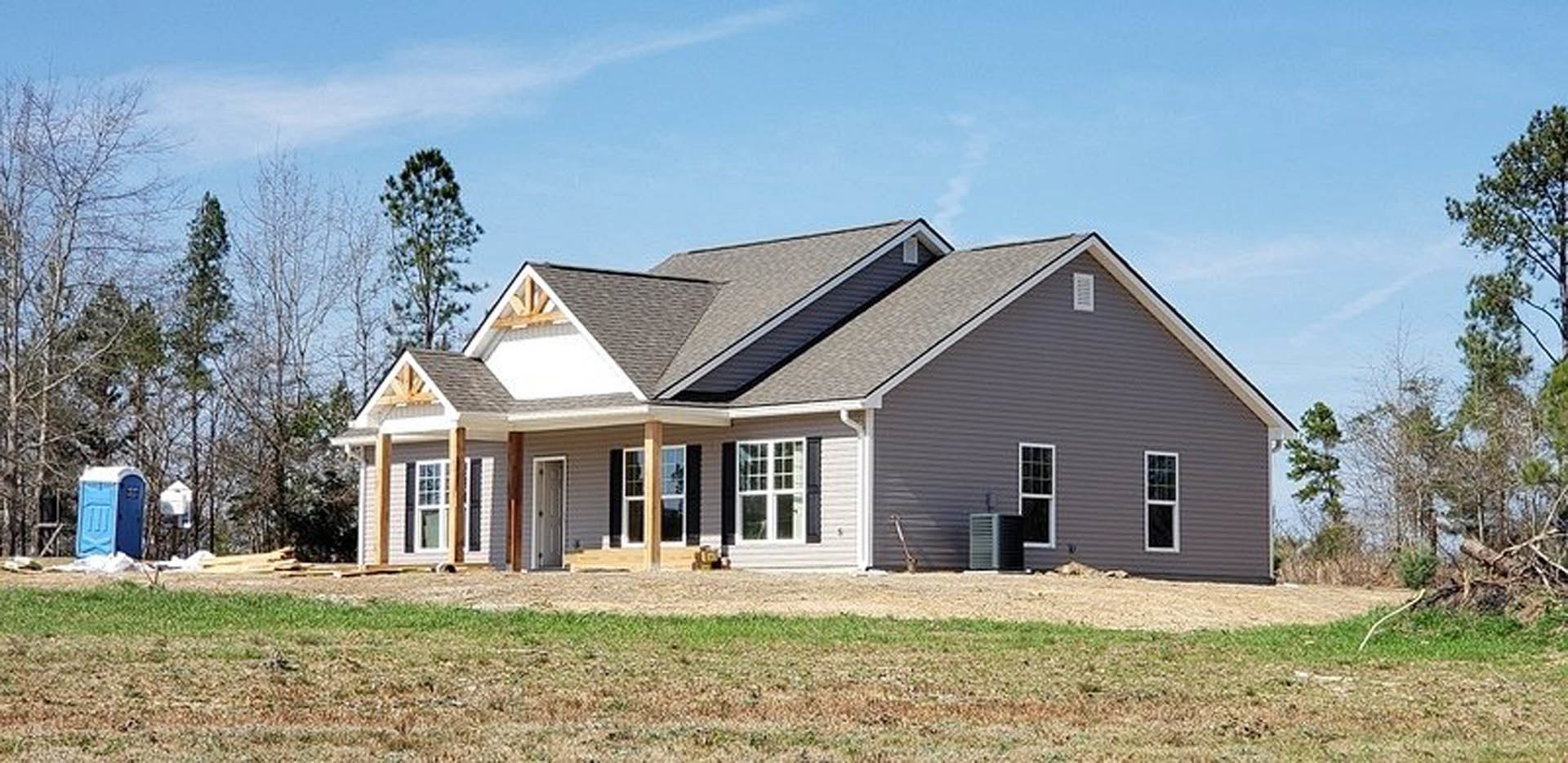 Partially built house with exposed framing, shingled roof, white framed windows, black door knob, and expansive green lawn under cloudy sky