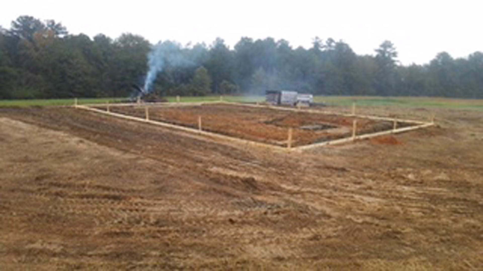 Square concrete foundation set in a grassy field, bordered by soil and sparse vegetation, with a distant fence and trees under a clear sky.