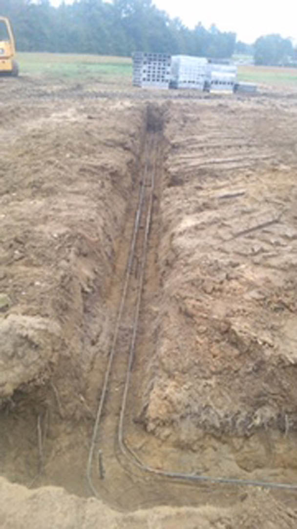 Open trench in muddy yard with exposed pipes and soil, construction equipment visible in background.