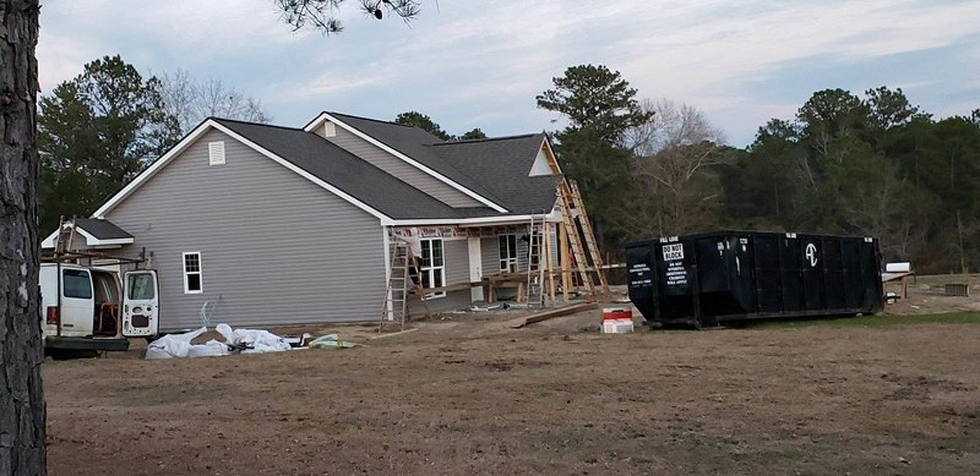 Partially built house with exposed framing, ladder leaning against exterior wall, black container on grassy area, open van door, dirt field with wooden beam, cloudy sky and trees