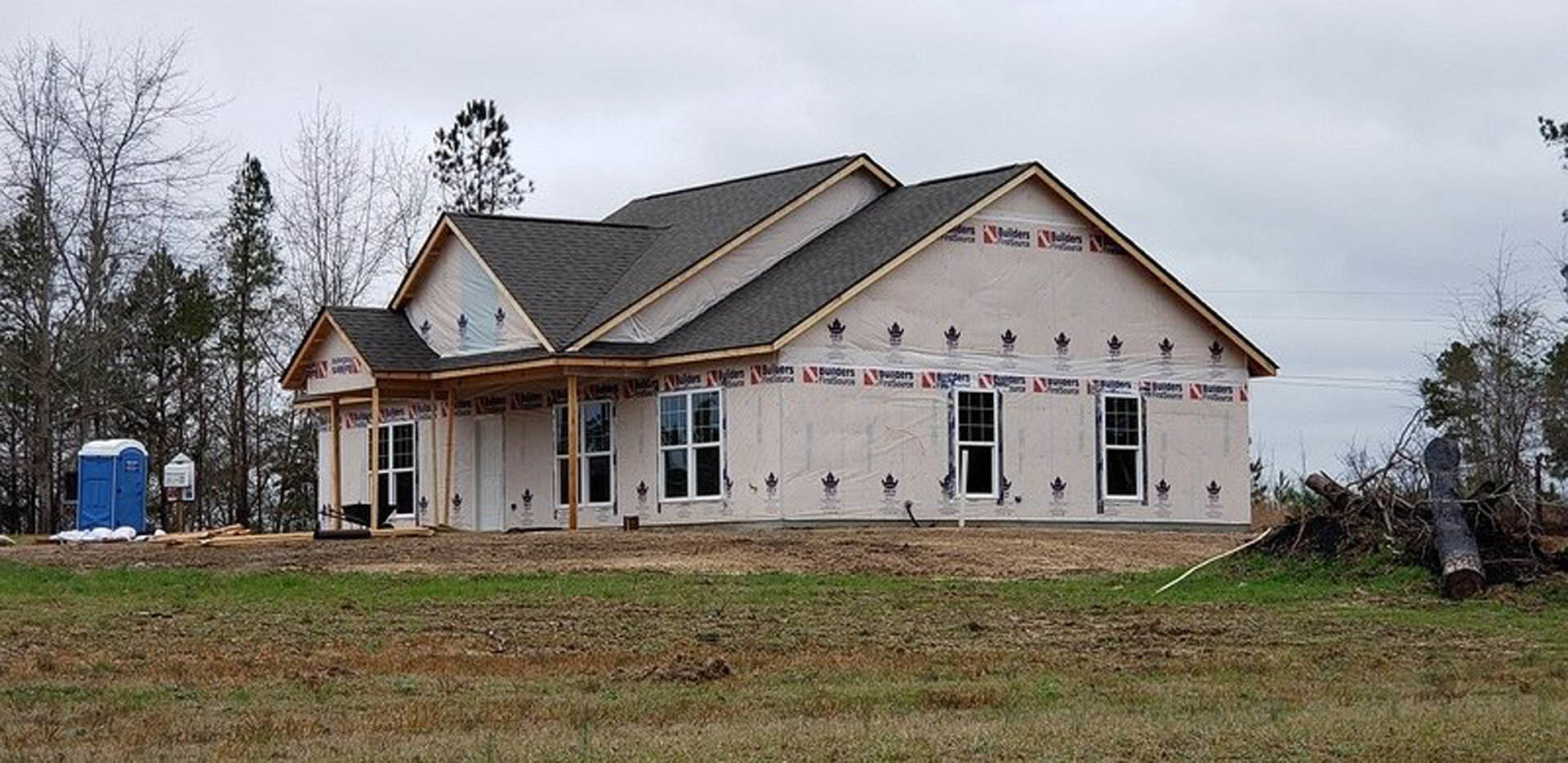 Partially built house with exposed framing, surrounded by a wooden fence, grassy yard, and scattered trees under a cloudy sky