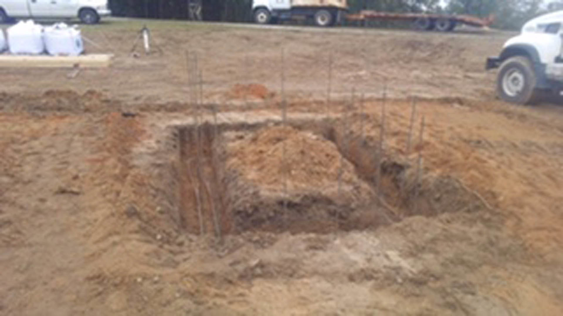 Excavated dirt pit in foreground with blurred white car and building in background