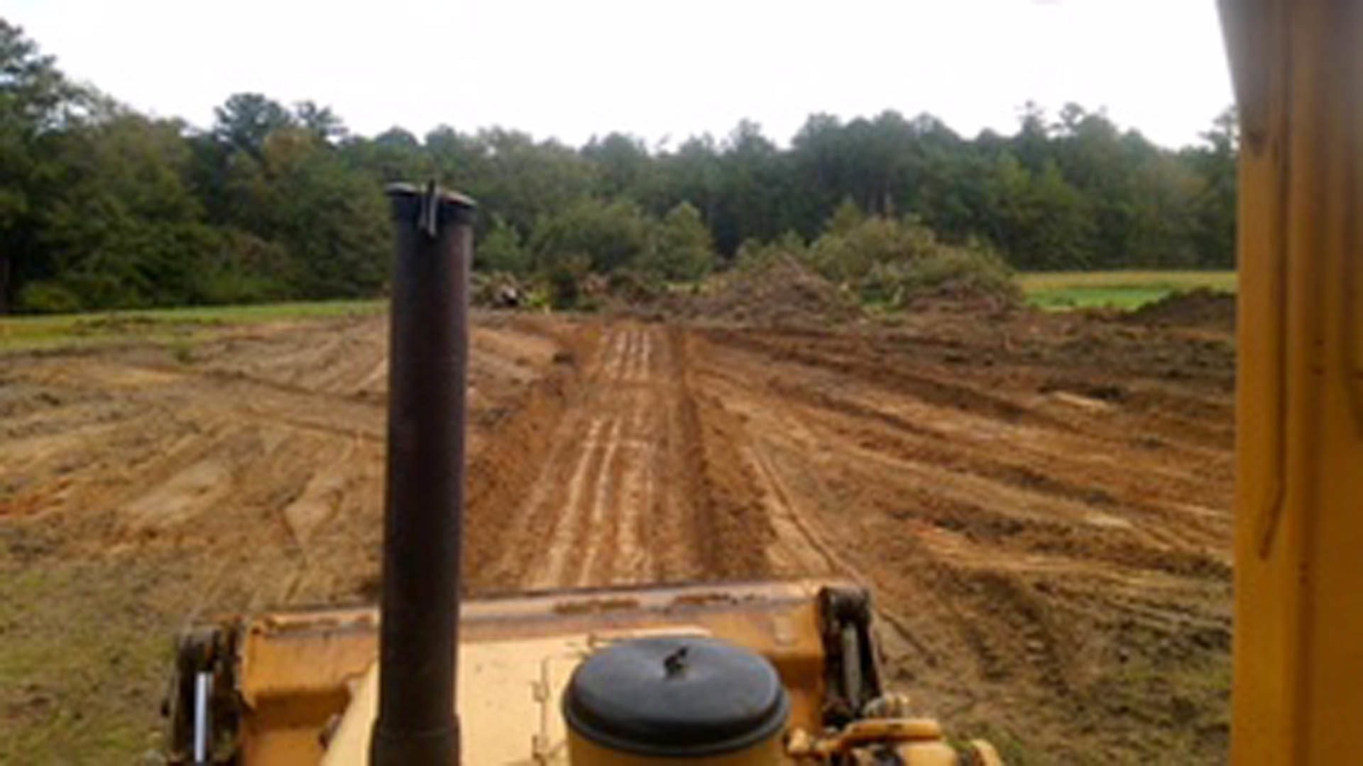 Modern farmhouse with wood siding and large windows, surrounded by trees and grass, tractor driving through dirt field in foreground