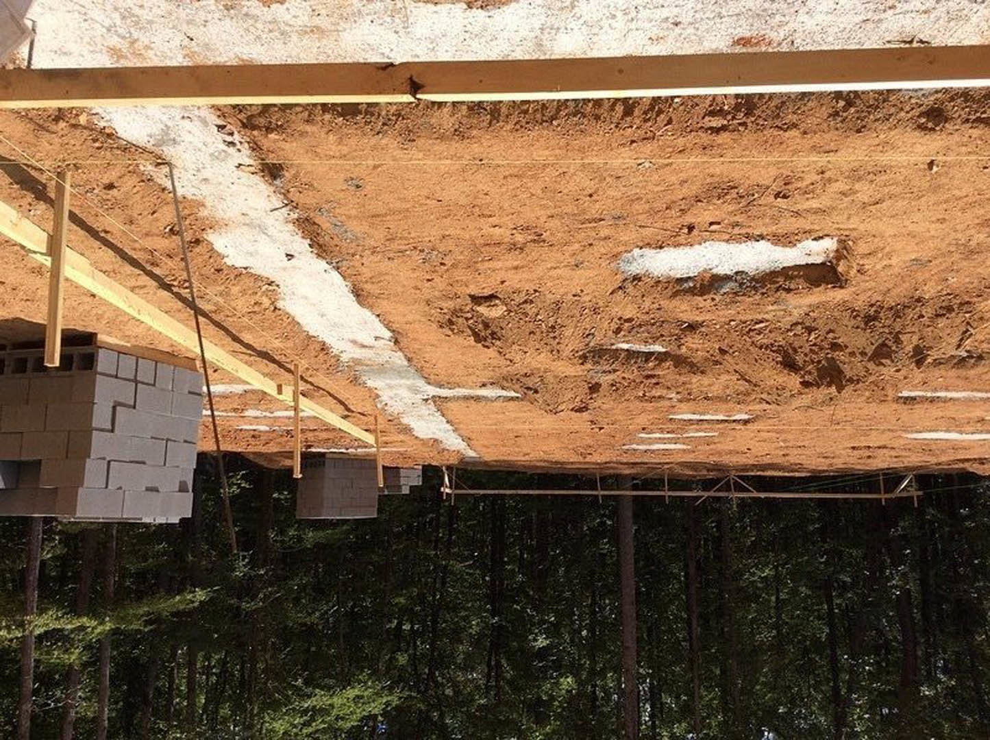 Stack of grey bricks and wooden beam on dirt construction site, partially built house with roof and trees in background, white substance scattered on ground