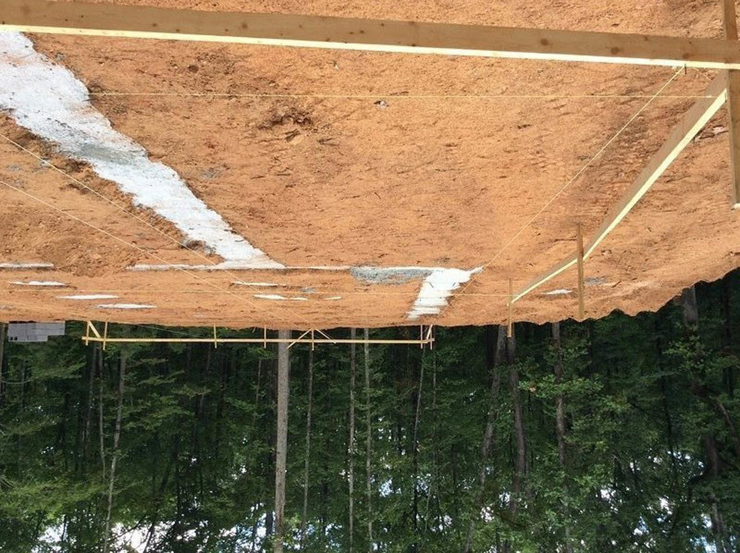 Dirt construction site bordered by trees, partially built wooden framing, white paint markings on ground, man standing beneath exposed beam