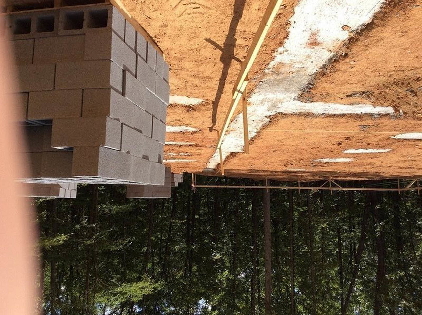Stack of red bricks on a dirt construction site beside a partially built wooden frame, with a finished house and trees in the background.