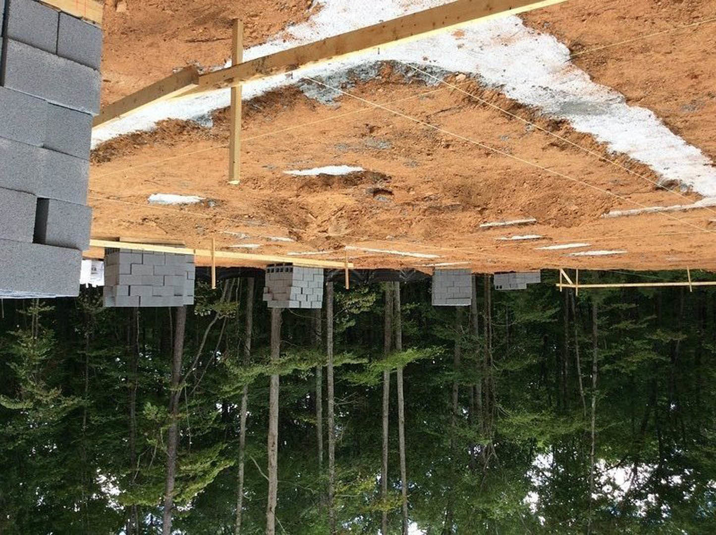Wood-framed custom home under construction with exposed brick chimney, grey block wall, and stacks of bricks surrounded by mature trees