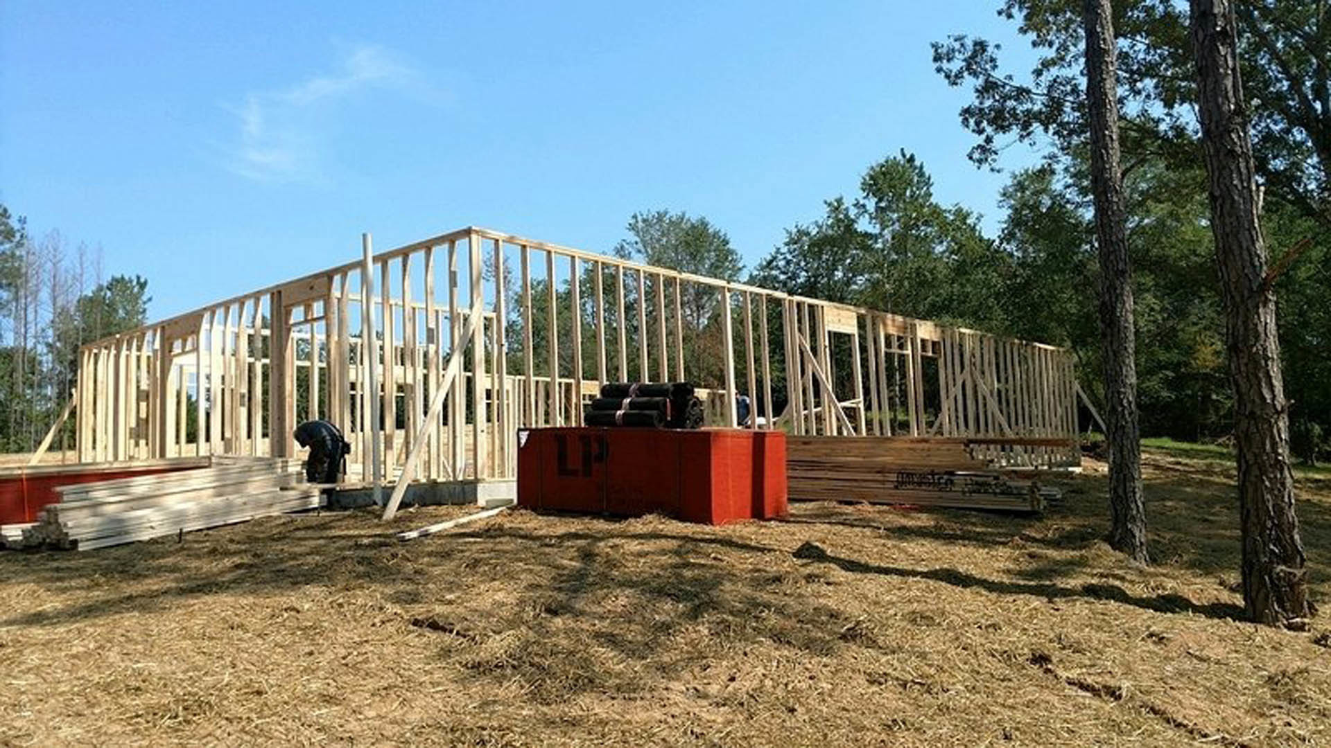 Wood-framed residential structure under construction, exposed beams and plywood, surrounded by dirt ground and temporary fencing, trees and sky in background
