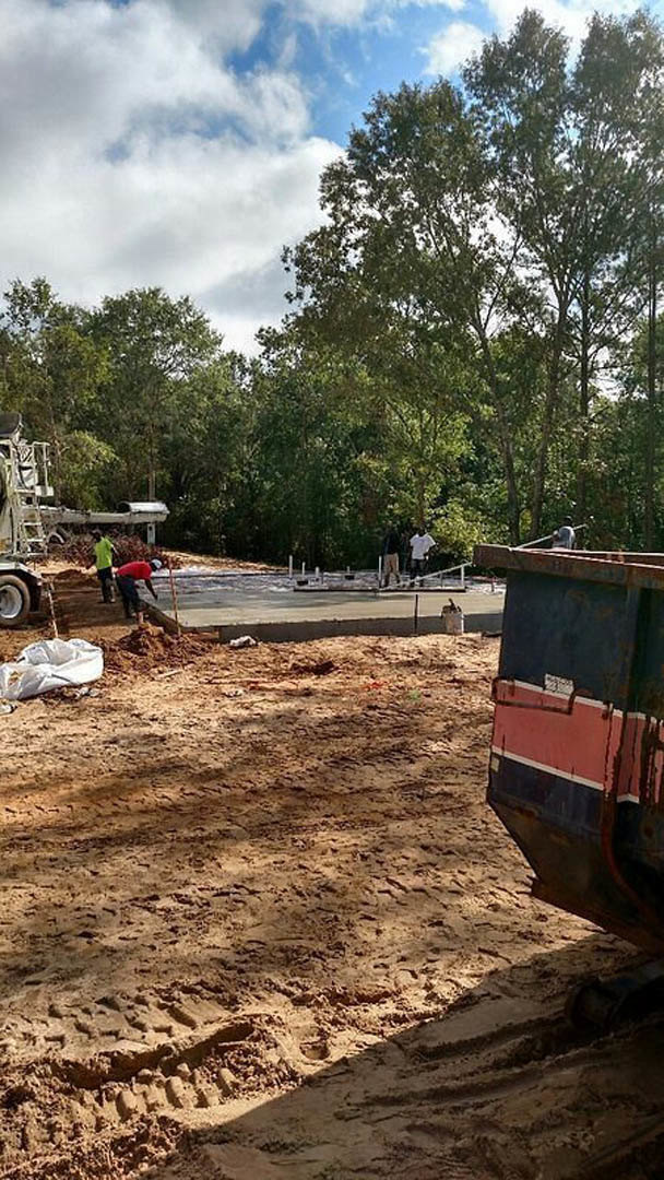 Framed custom home under construction with exposed wood beams, sandy soil marked by tire tracks, construction truck parked nearby, workers on site, surrounded by mature trees and