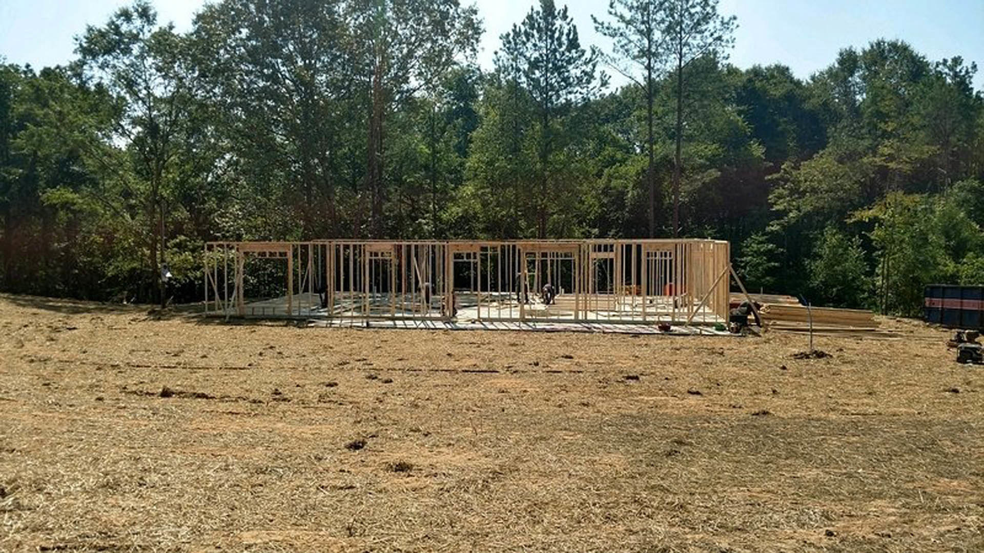 Wood-framed custom home under construction on dirt lot, surrounded by grass and trees, with exposed beams and unfinished exterior walls.