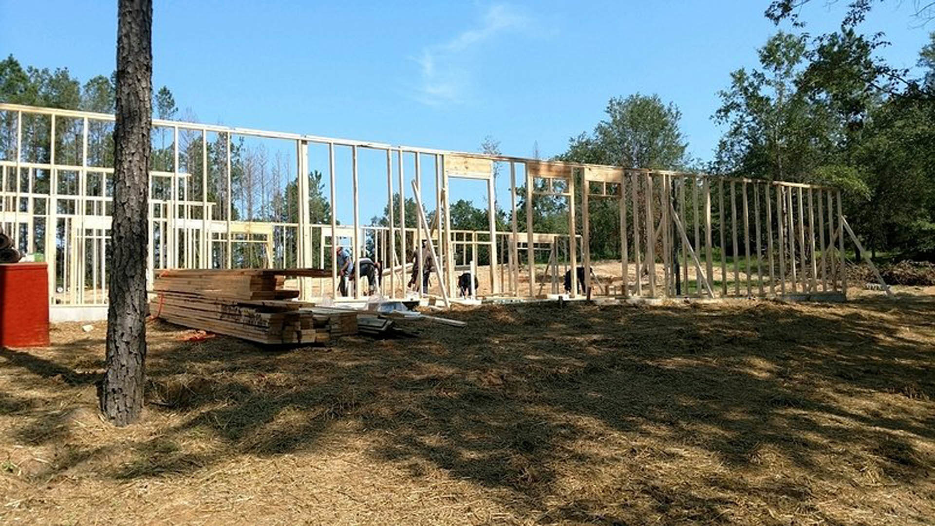 Wood-framed custom home under construction with exposed beams, surrounded by grass, trees, and a fenced yard under a clear sky