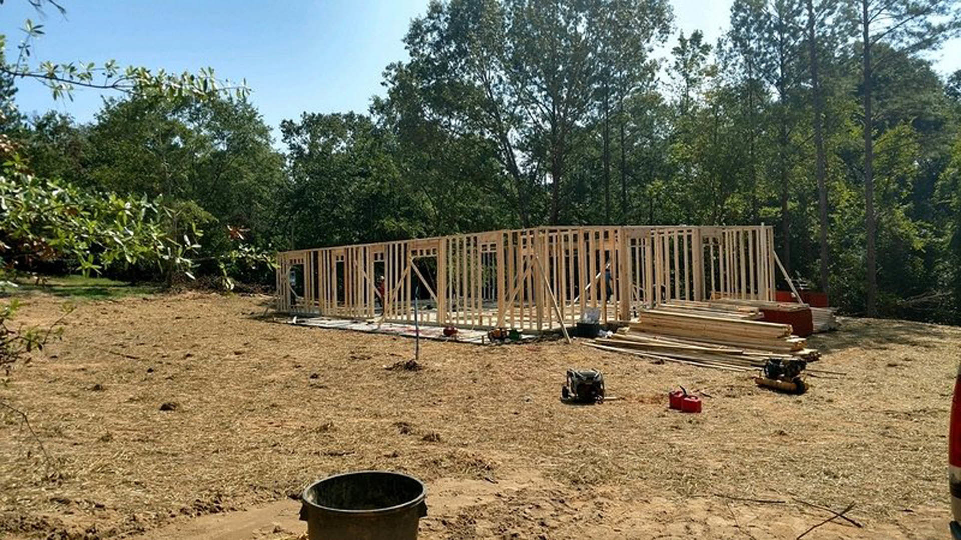 Framed custom home under construction with exposed wood beams, unfinished exterior walls, and surrounding mature trees in the background