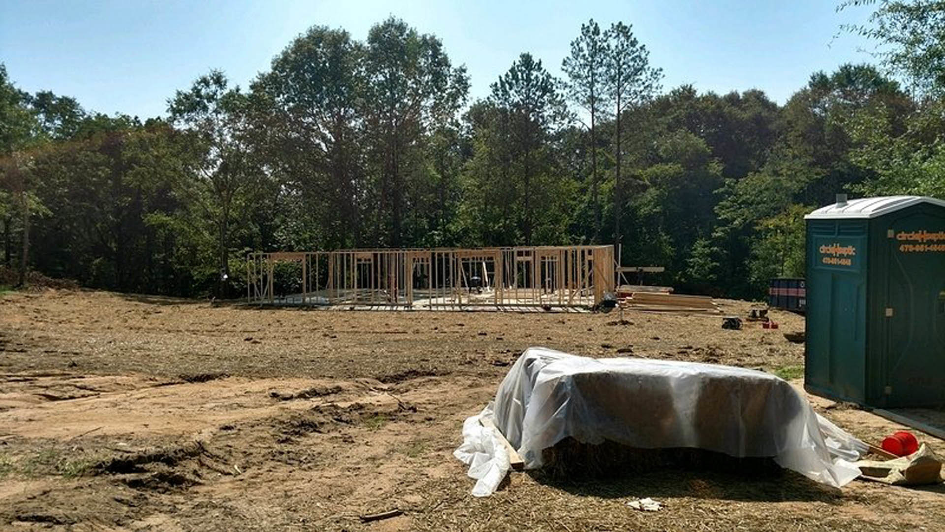 Wood-framed house under construction with exposed beams, green portable toilet, white plastic-covered table, hay bale wrapped in plastic, trees in background, soil and construction