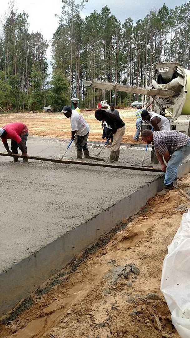Workers pouring and leveling concrete slab outdoors, surrounded by soil and trees, with men holding shovels and metal rods, some wearing red shirts and white tank tops.