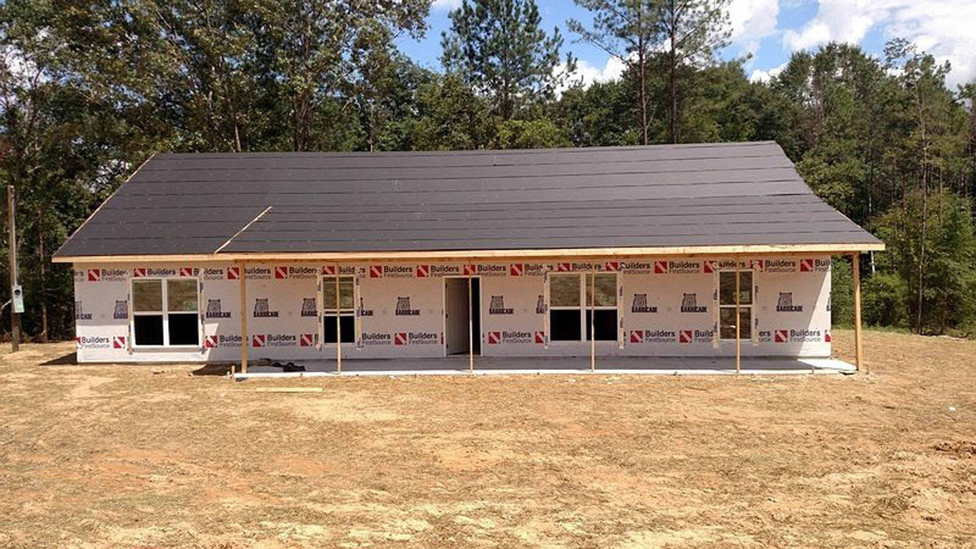 Partially built house with exposed wooden framing, grey roof, large windows, and surrounding trees in the background