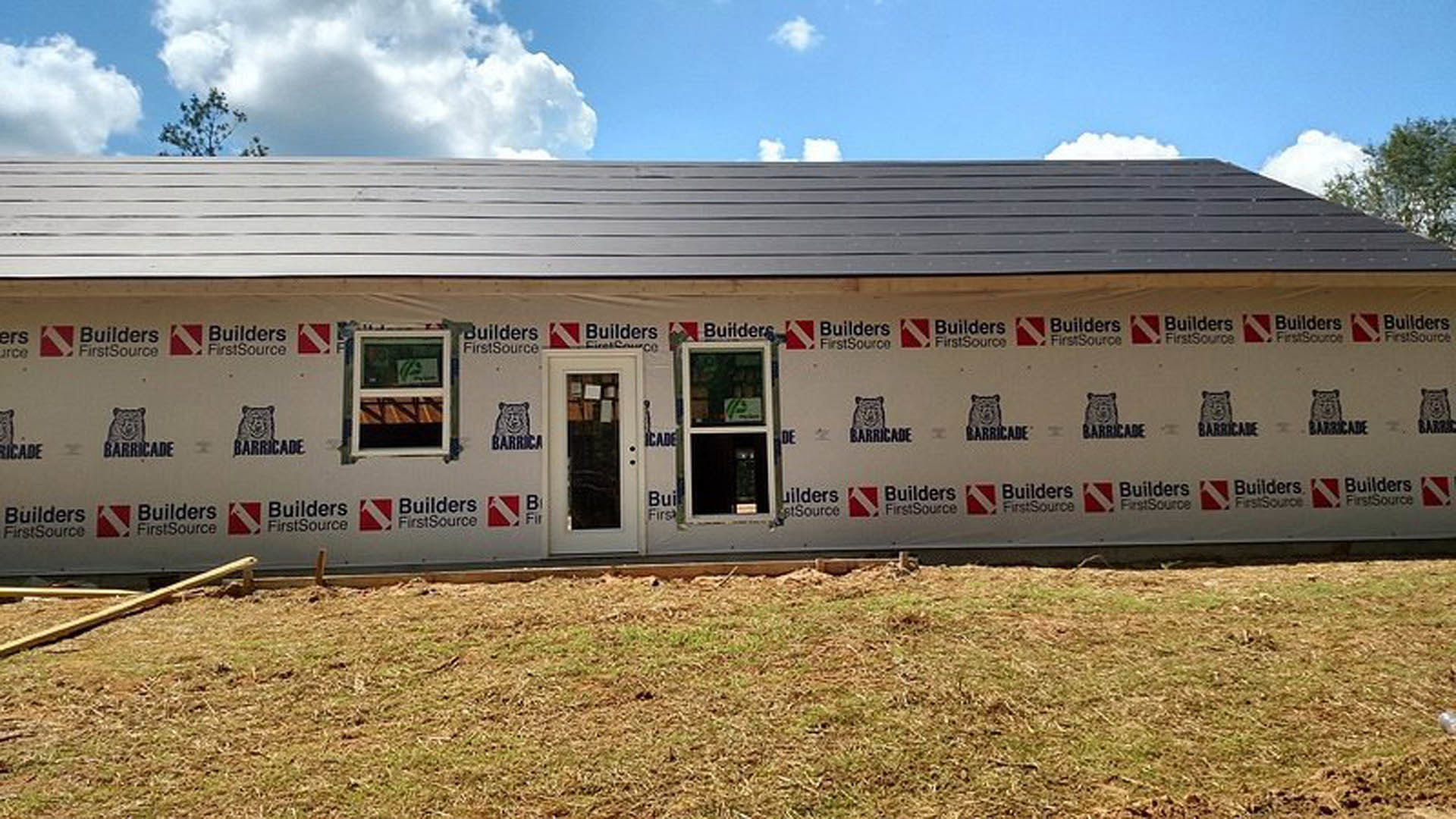 Modern home under construction with unfinished exterior walls, glass door, large window, and patch of green grass in front; bear face sign visible near entrance.