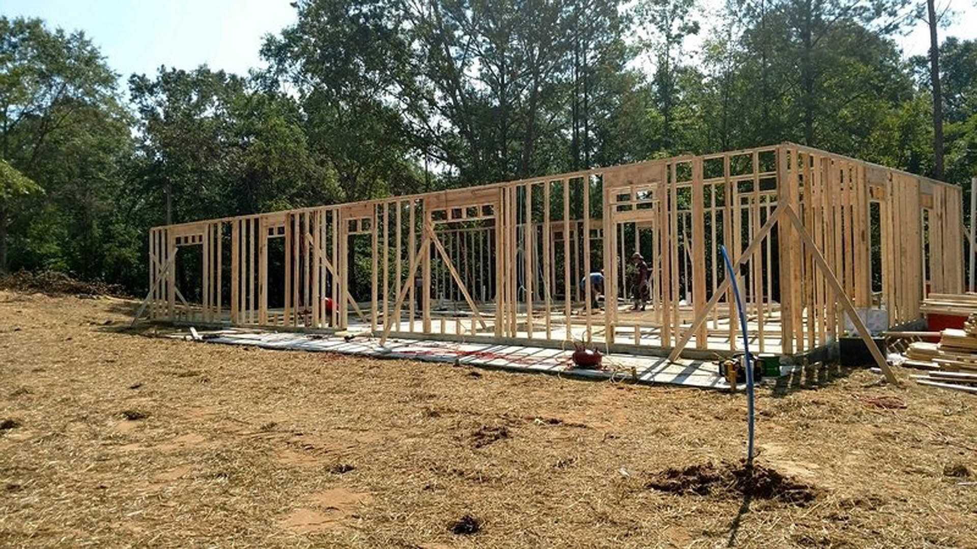 Wood-framed custom home under construction with exposed foundation, unfinished walls, and surrounding trees in the background