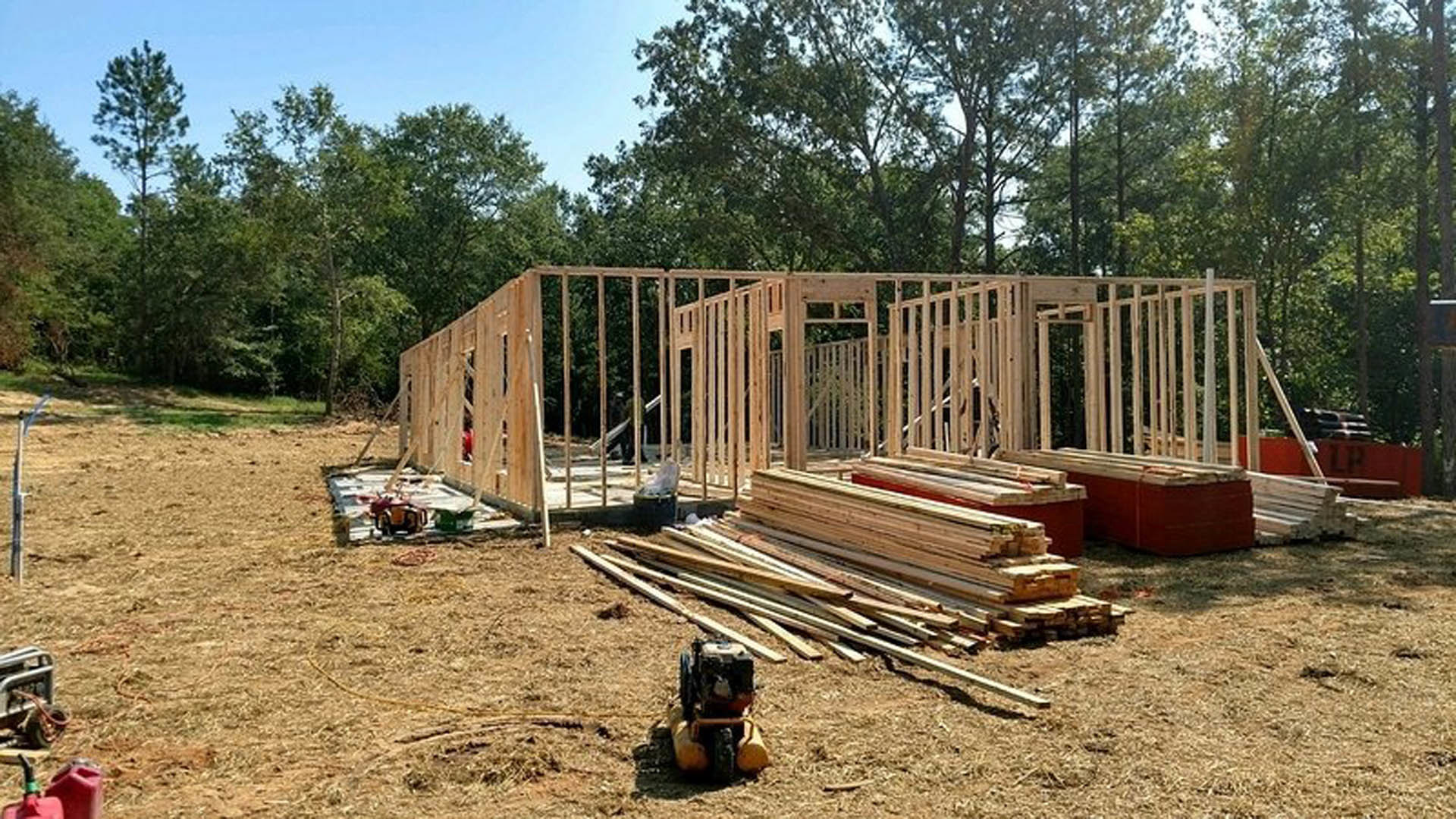 Wood-framed custom home under construction with exposed lumber, foundation, and stacks of wood planks on dirt ground; trees and blue sky in background