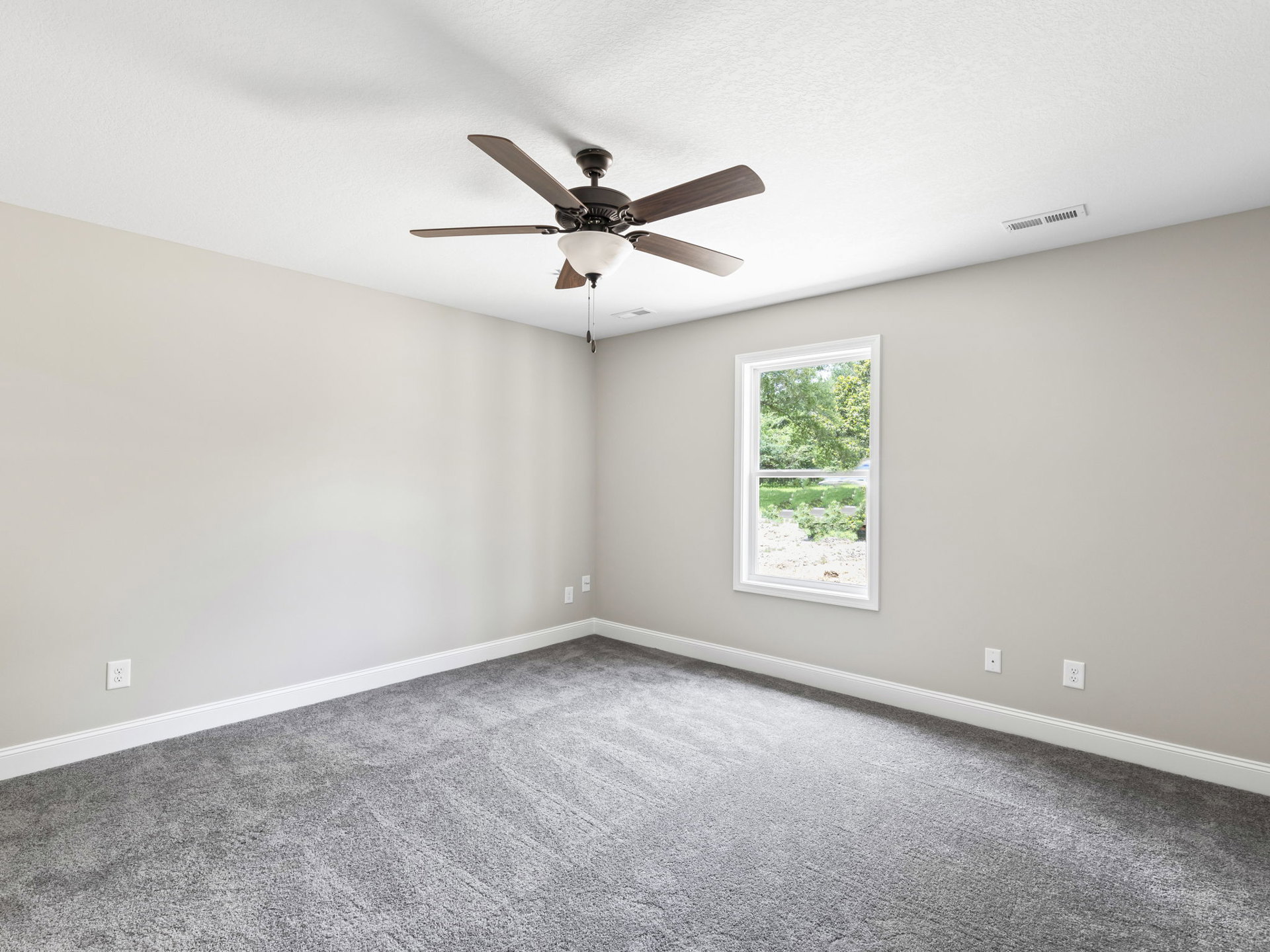 Neutral-toned room with grey carpet, white walls, ceiling fan with light fixture, and window overlooking leafy trees
