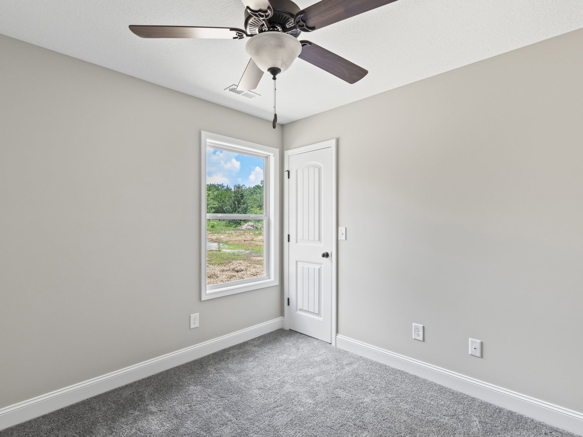 Ceiling fan with light fixture mounted on white plaster ceiling, carpeted floor, white door, and large window revealing trees and blue sky outside