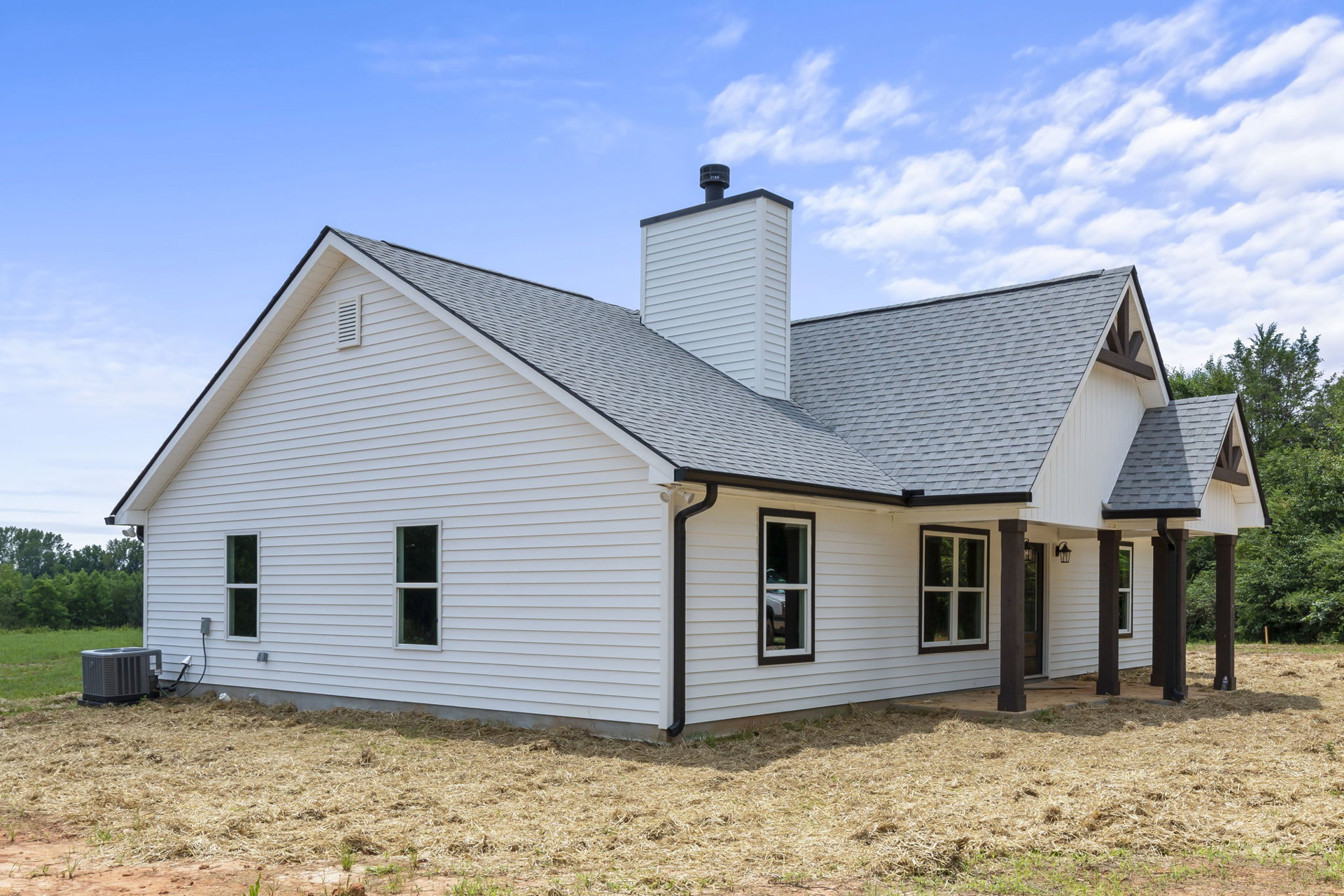 White siding house under construction beneath blue sky, visible chimney, white-framed windows including one reflecting a person, grey heat pump unit outside, partial view of nearby